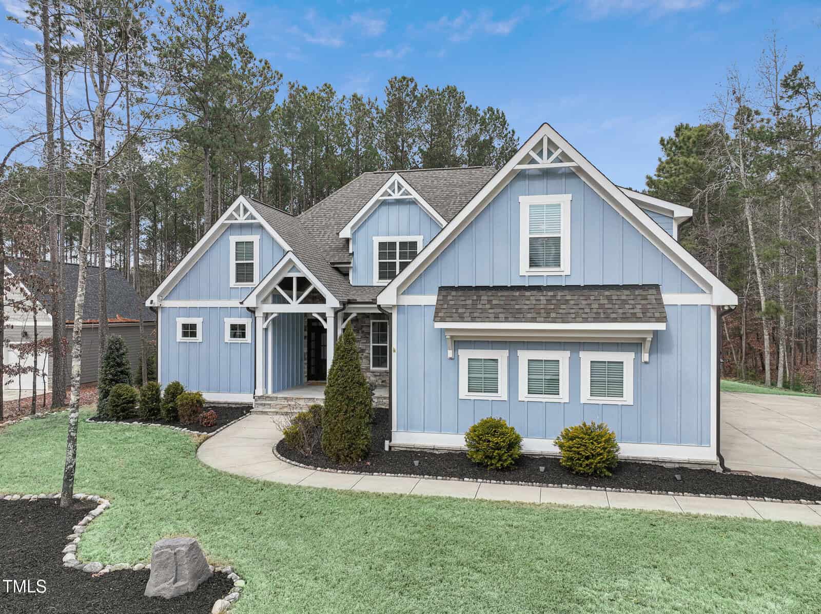 A blue two-story house with white trim and gabled roof is surrounded by a grass lawn and small shrubs. There is a driveway on the right side and trees in the background under a clear blue sky.