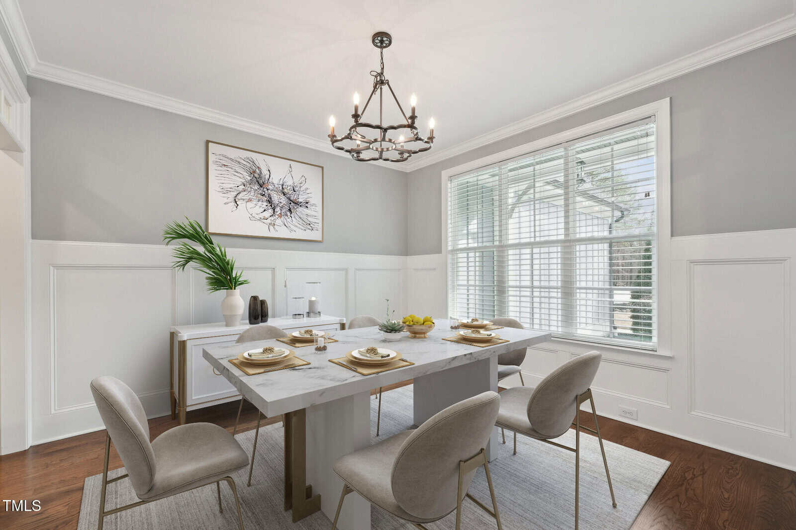 A dining room with a rectangular marble table set for four, featuring beige chairs. A large window with blinds allows natural light in. A chandelier hangs overhead. Walls are adorned with white wainscoting and gray paint, with a framed abstract art piece.