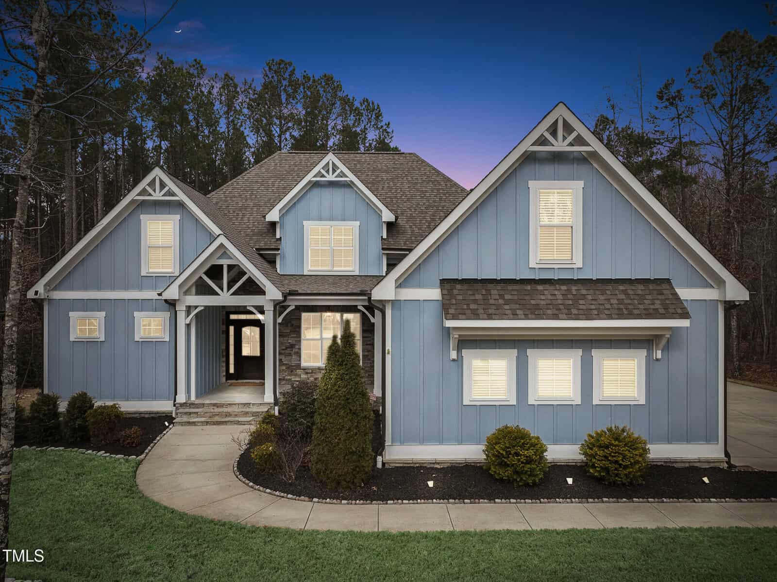 A large blue house with white trim and a complex roof design is set against a backdrop of trees. The home features multiple gables and a central entryway with stone steps. The sky is a gradient of blue and purple, suggesting twilight.