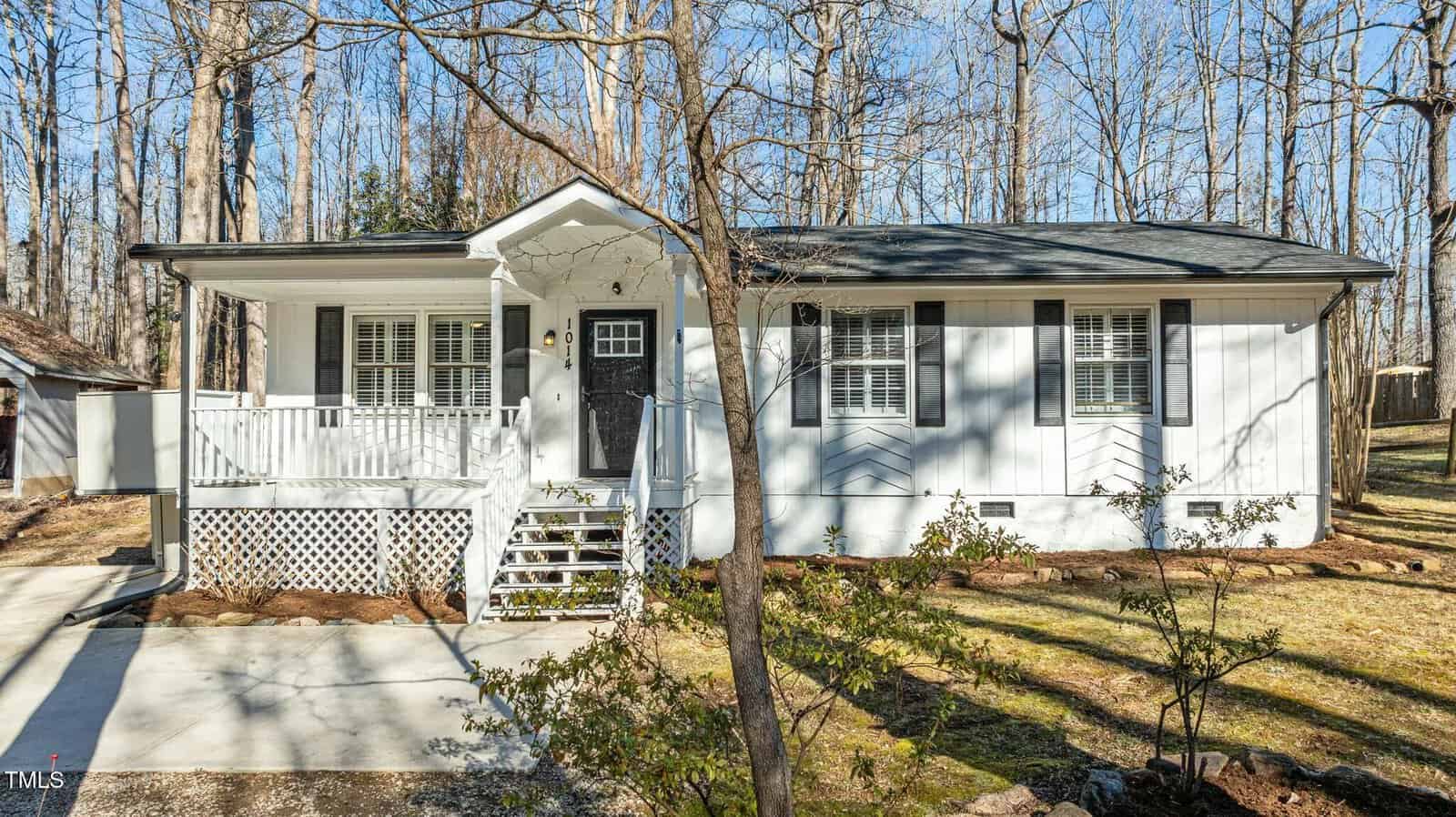 A single-story white house with black shutters and a covered front porch, surrounded by leafless trees and a grassy yard. A concrete driveway runs along the left side of the house.