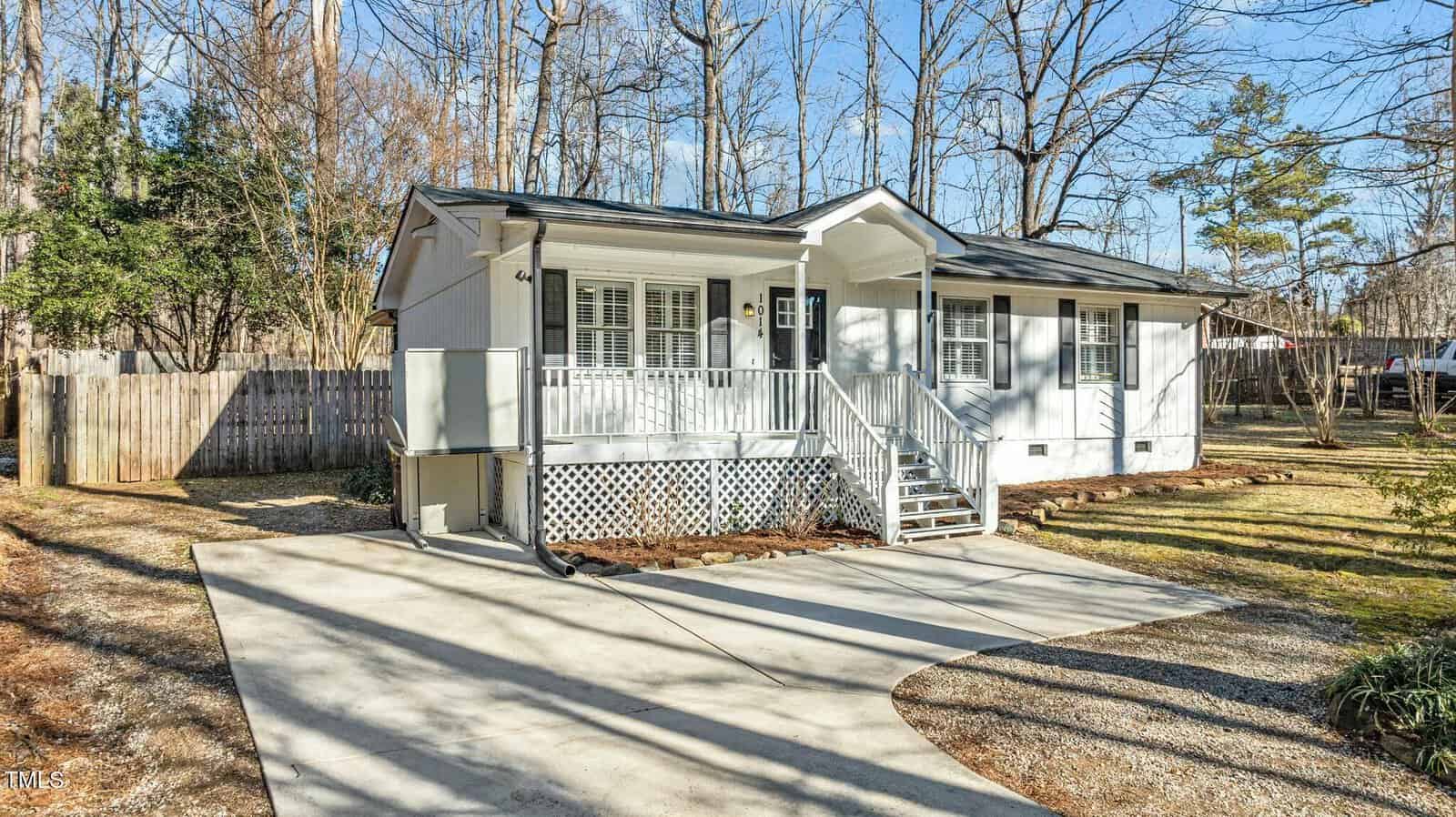 A small white house with a front porch, steps, and railing, sits in a wooded yard. There’s a driveway, fenced backyard, and leafless trees in the background under a clear blue sky.