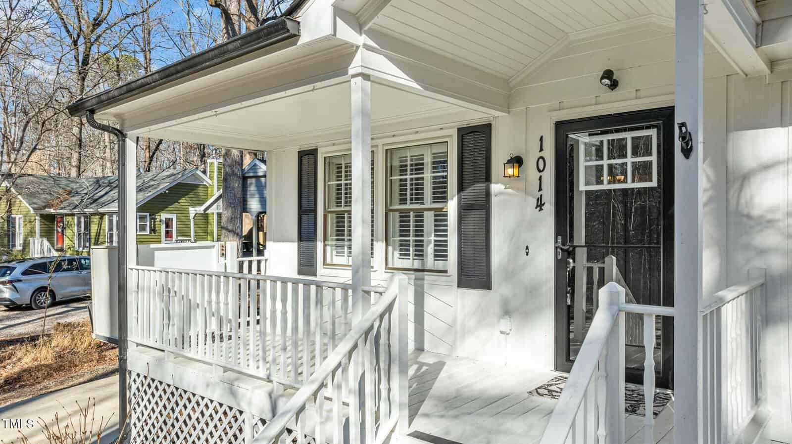 A white front porch with railing and steps leads to a black front door with glass panes. The house number "1011" is displayed next to the door. Neighboring homes and cars are visible in the background.