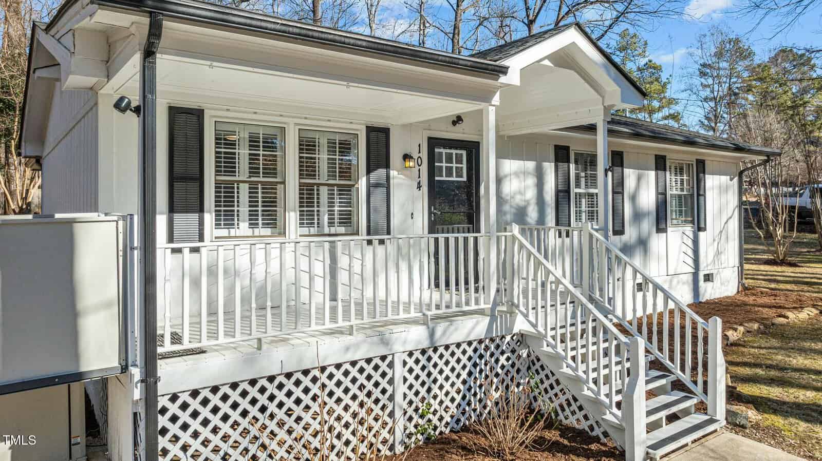 White single-story house with a covered front porch, black shutters, white railing, and steps. Surrounded by trees and a well-maintained yard on a sunny day.