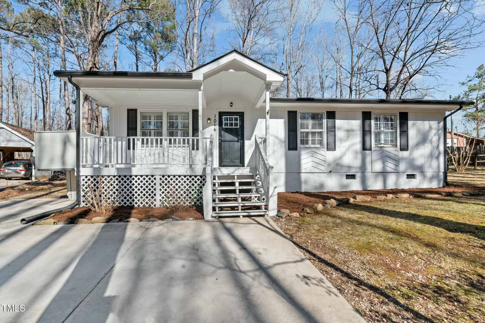 A single-story white house with a front porch, gray shutters, and a central door. Steps lead up from a concrete driveway. Leafless trees surround the house, and the yard has patches of grass and mulch.