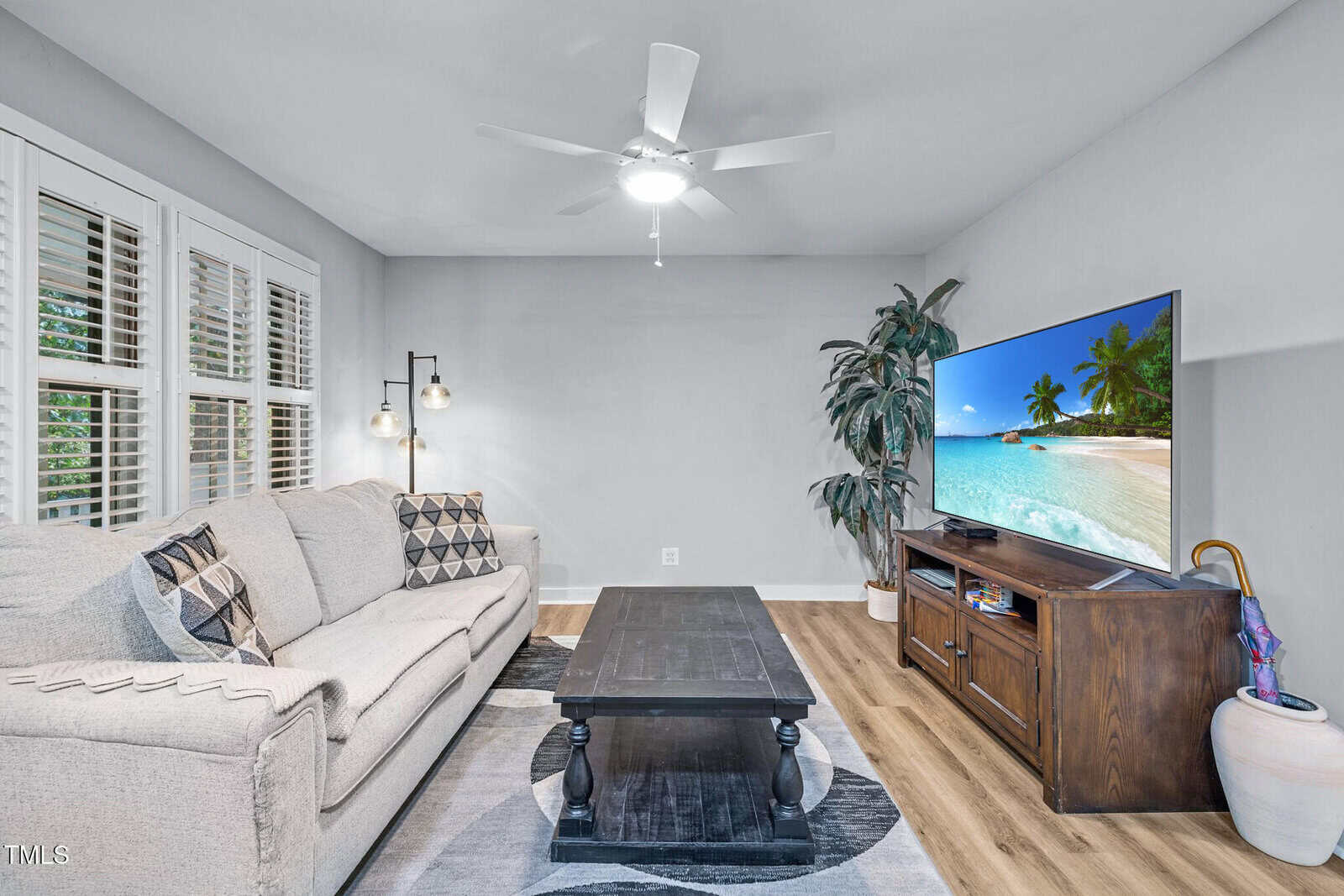 A living room with a light gray sofa, patterned cushions, a black coffee table, a TV displaying a beach scene, a large plant, wood flooring, white shutters, and a ceiling fan with a light.