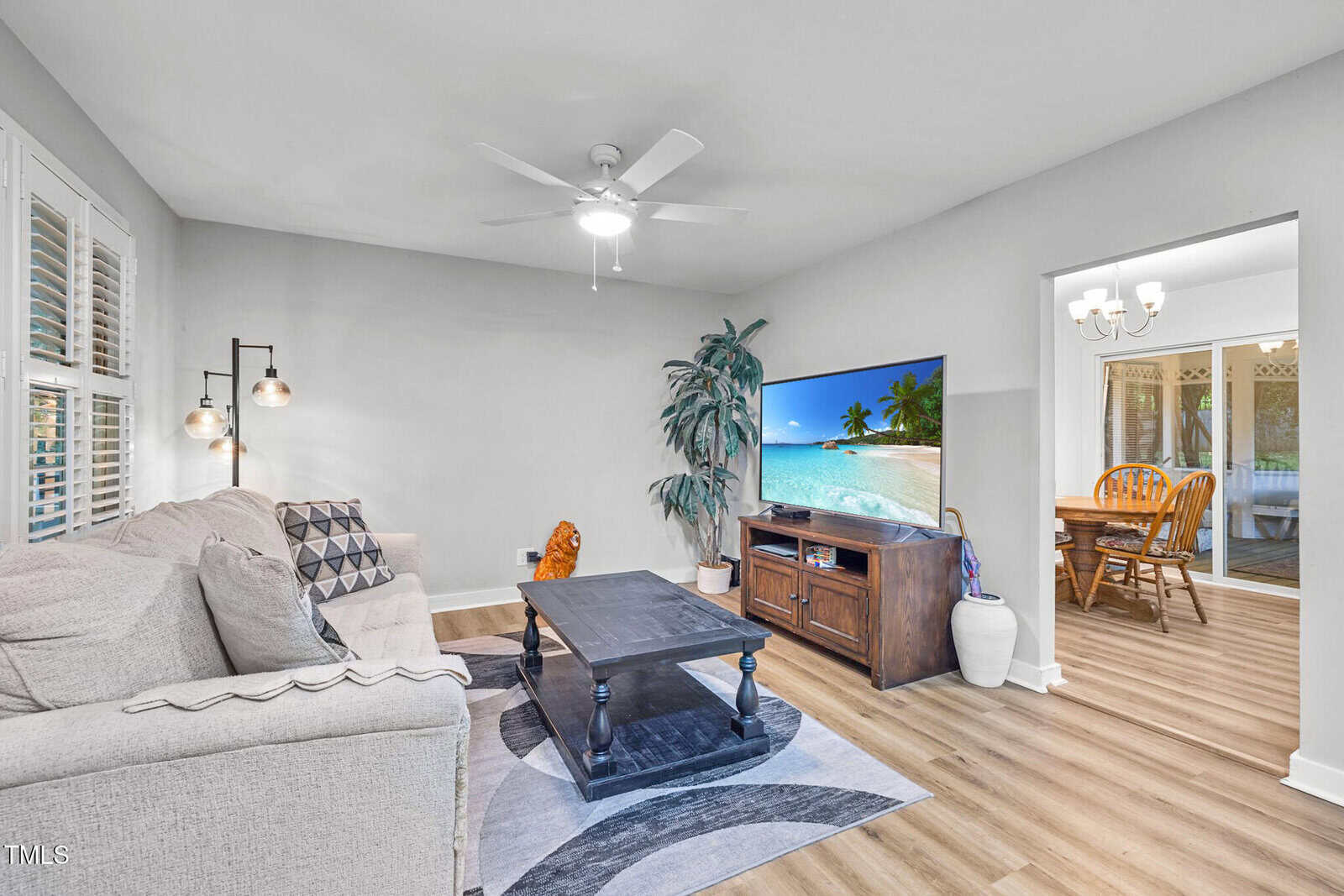 A living room with light gray walls, a beige sectional sofa, a dark wood coffee table, a TV displaying a beach scene, and a large plant. A dining area with a wooden table and chairs is visible through an open doorway.