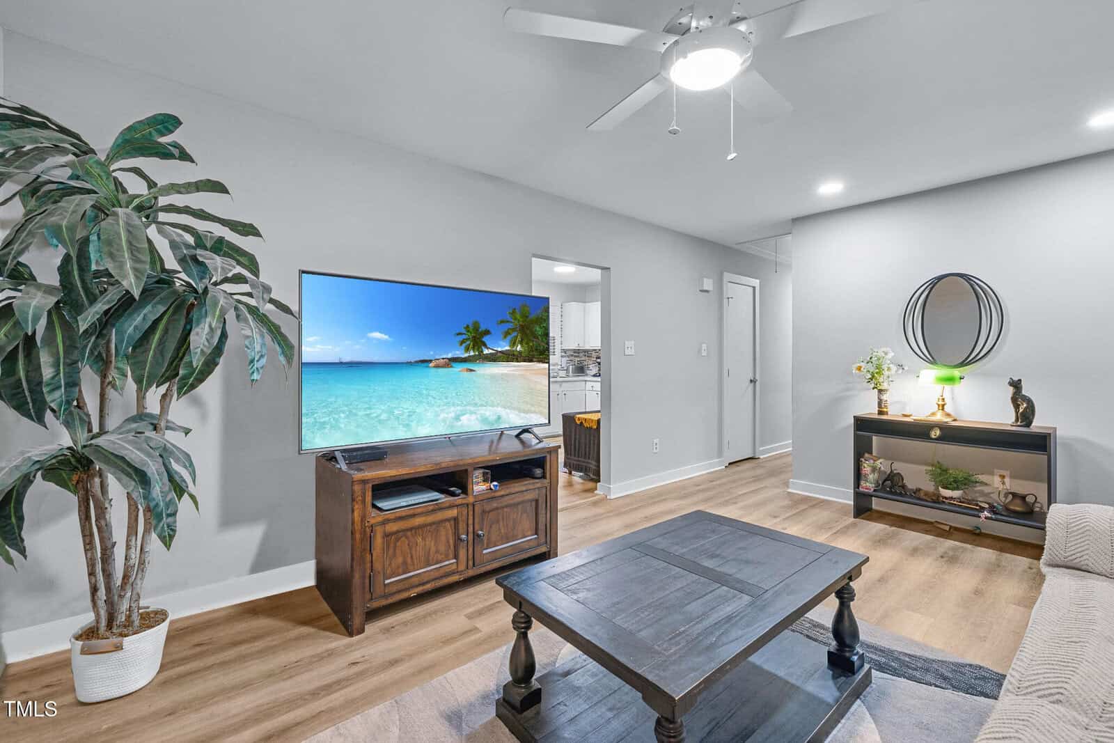 A modern living room with a ceiling fan, TV displaying a beach scene, a wooden coffee table, a console table with decor, a potted plant, and wood flooring. Walls are light gray and lighting is recessed.