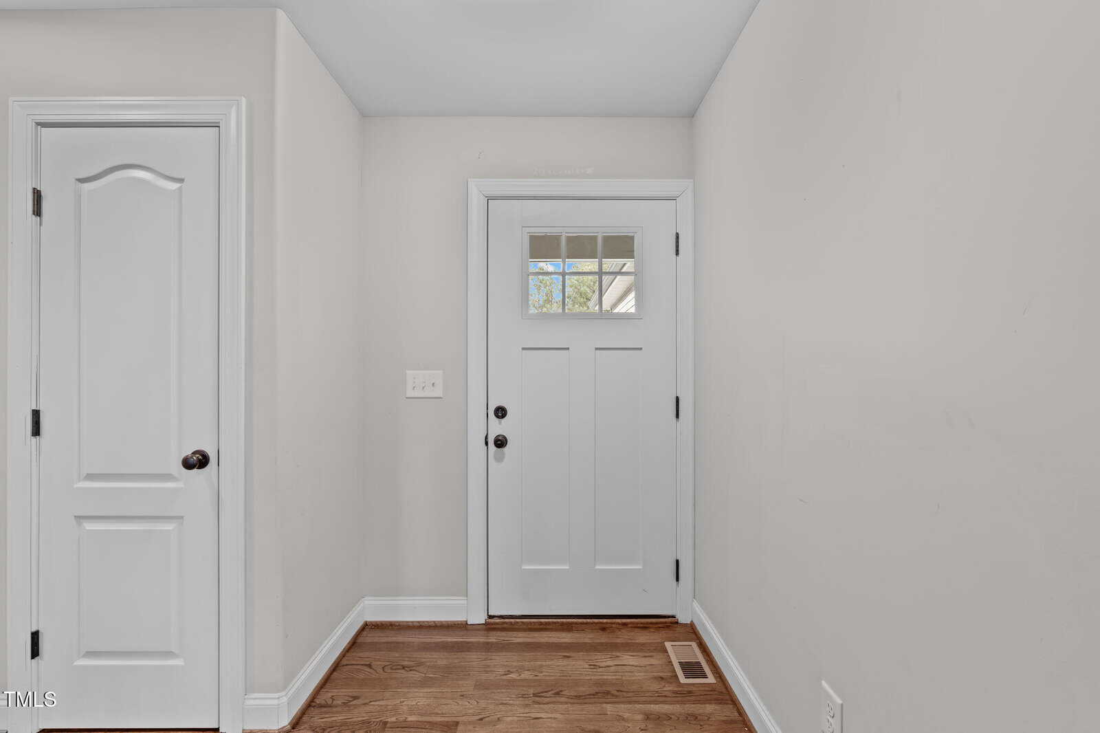 A small hallway featuring a light gray front door with a windowpane and a white interior door on the left. The floor is made of wooden planks, and there is a light switch on the wall.