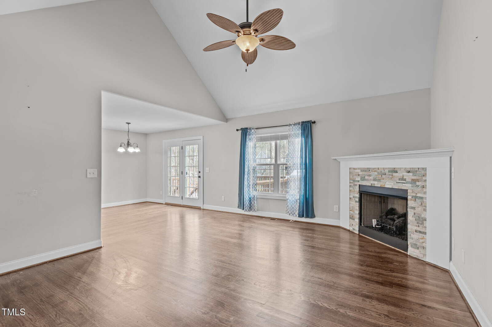 A spacious room with light gray walls and a wooden floor. It features a ceiling fan with a light in the center, an empty fireplace with a tiled surround, and windows with sheer blue curtains. Double glass doors open to an outdoor deck area.