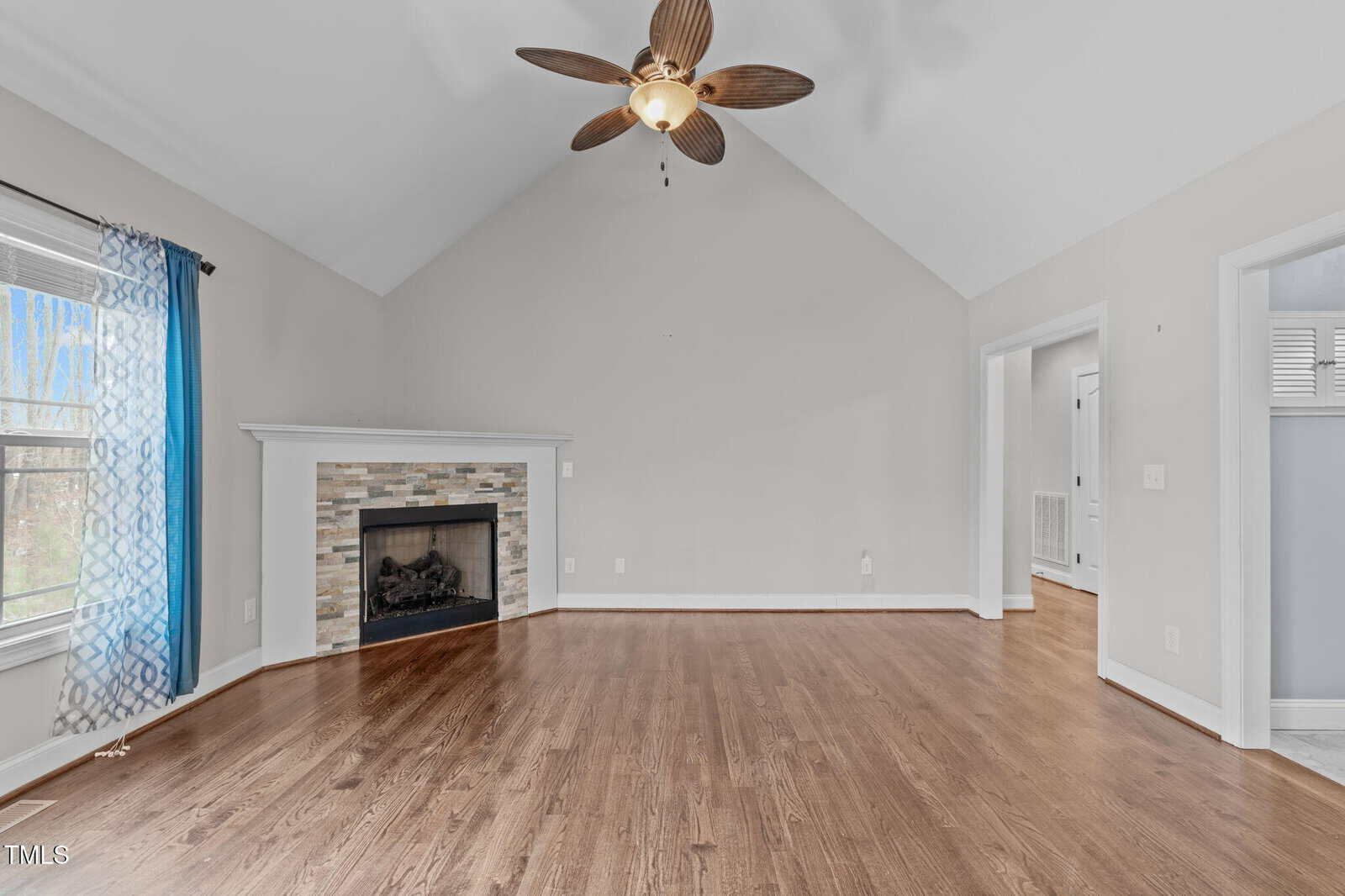 A spacious, unfurnished room with wooden floors and light-colored walls. The room features a fireplace with a stone surround, a ceiling fan with wooden blades, and a window with a blue curtain. The ceiling is vaulted.