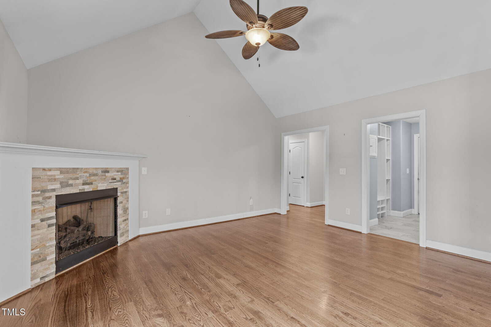 A spacious room with polished hardwood floors, a large stone-framed fireplace, and a vaulted ceiling. A ceiling fan with a light hangs above. There are two doorways leading to other rooms with light-colored walls.