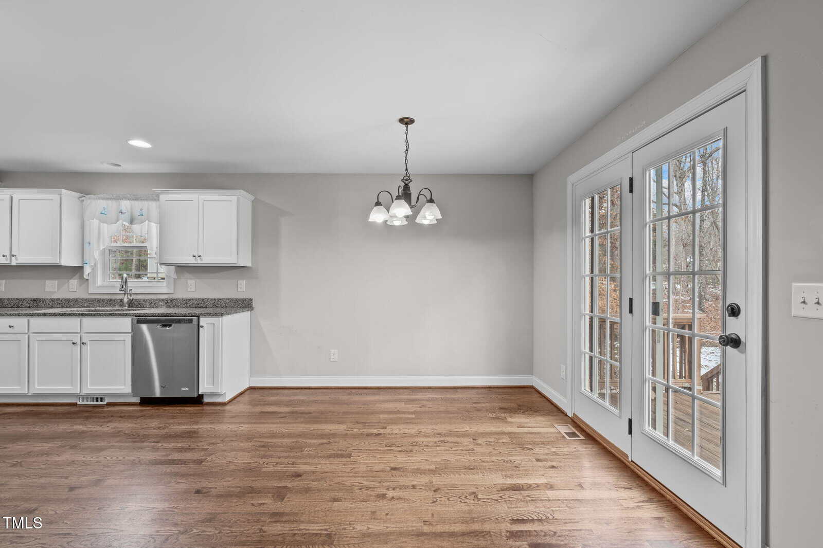 Bright kitchen and dining area with hardwood floors. White cabinets and stainless steel dishwasher are on the left. A hanging light fixture is centered above the dining space. Double glass doors lead to an outdoor area on the right.