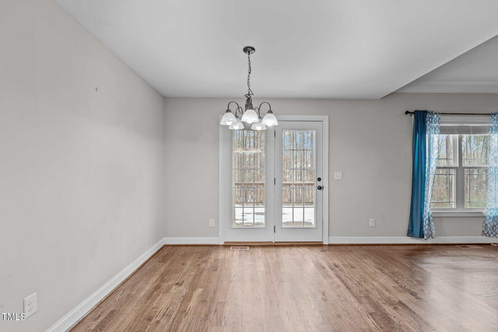 A room with wooden flooring and a hanging chandelier. There is a glass door leading outside and a window with blue curtains. Walls are painted light gray, and the room appears empty. Natural light is coming in through the glass door and window.