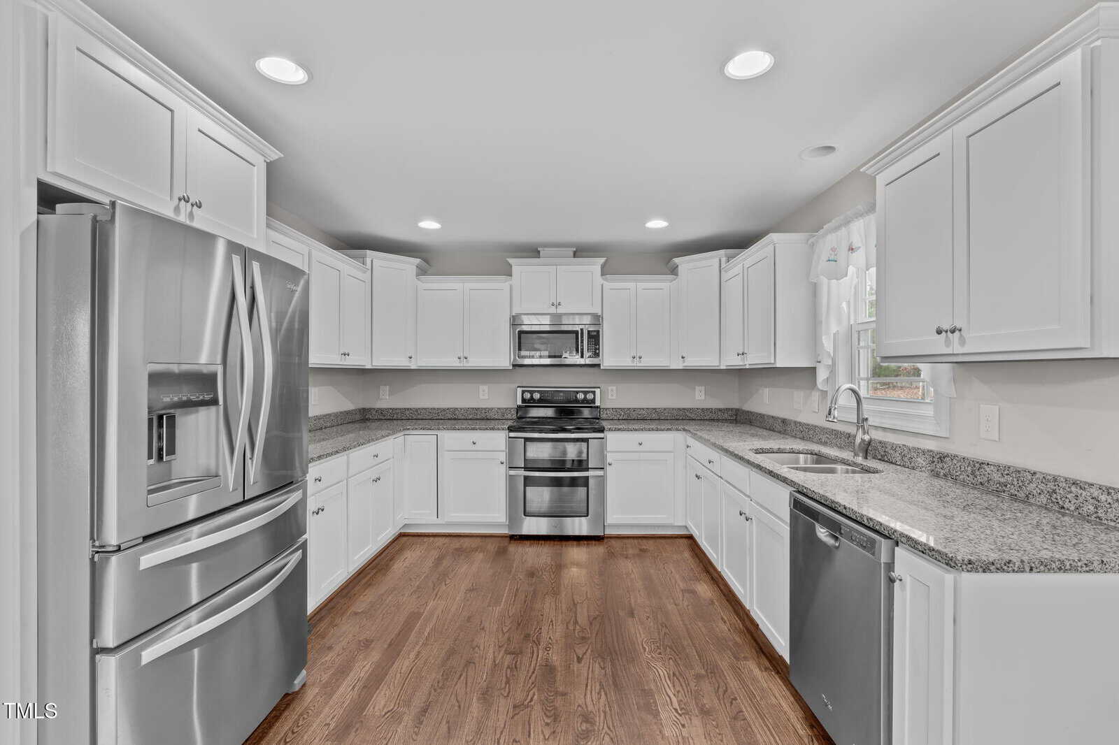 A modern kitchen with white cabinets, stainless steel appliances including a double oven, refrigerator, and dishwasher. Granite countertops line the walls, and the floor is made of wood. Recessed lighting illuminates the space.