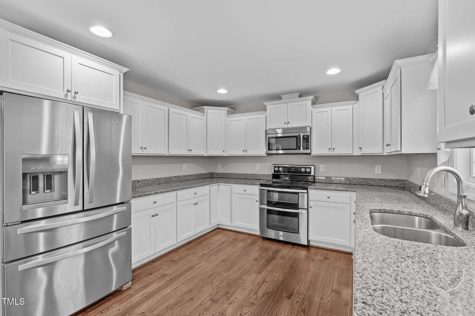A modern kitchen with stainless steel appliances, including a refrigerator, stove, and microwave. White cabinetry lines the walls, and a granite countertop includes a sink with a curved faucet. The floor is made of polished wood.