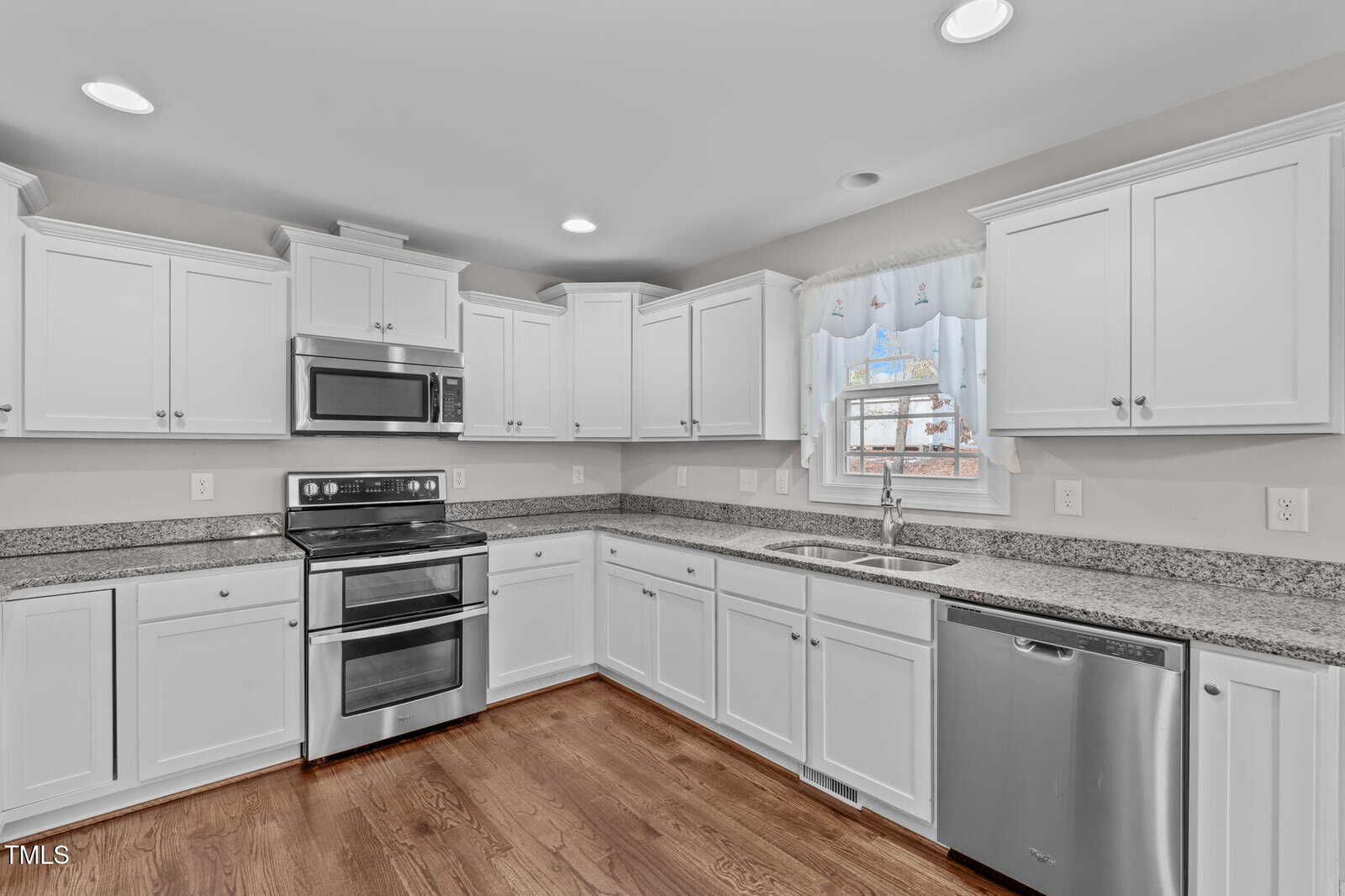 A modern kitchen with white cabinets, stainless steel appliances including a double oven and dishwasher, and granite countertops. A window above the sink has a decorative curtain. The floor is wooden, and recessed lighting is in the ceiling.