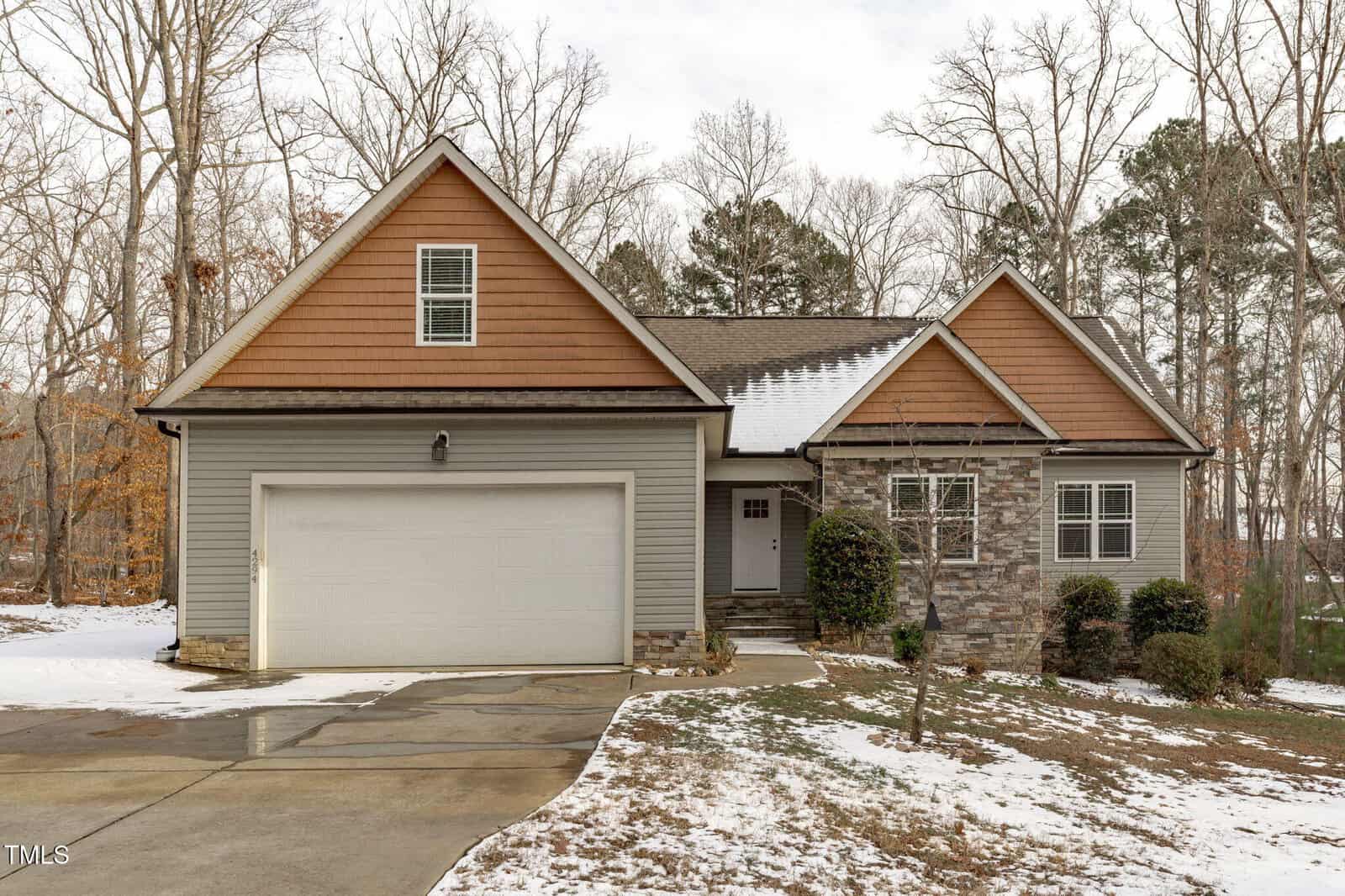 This charming single-story house at 4294 Sustain Circle, Franklinton, boasts gray siding with brick accents. It features a two-car garage and a small porch. The roof, covered in shingles, is framed by bare trees. A light snow dusts the driveway, flanked by shrubs near the entrance.