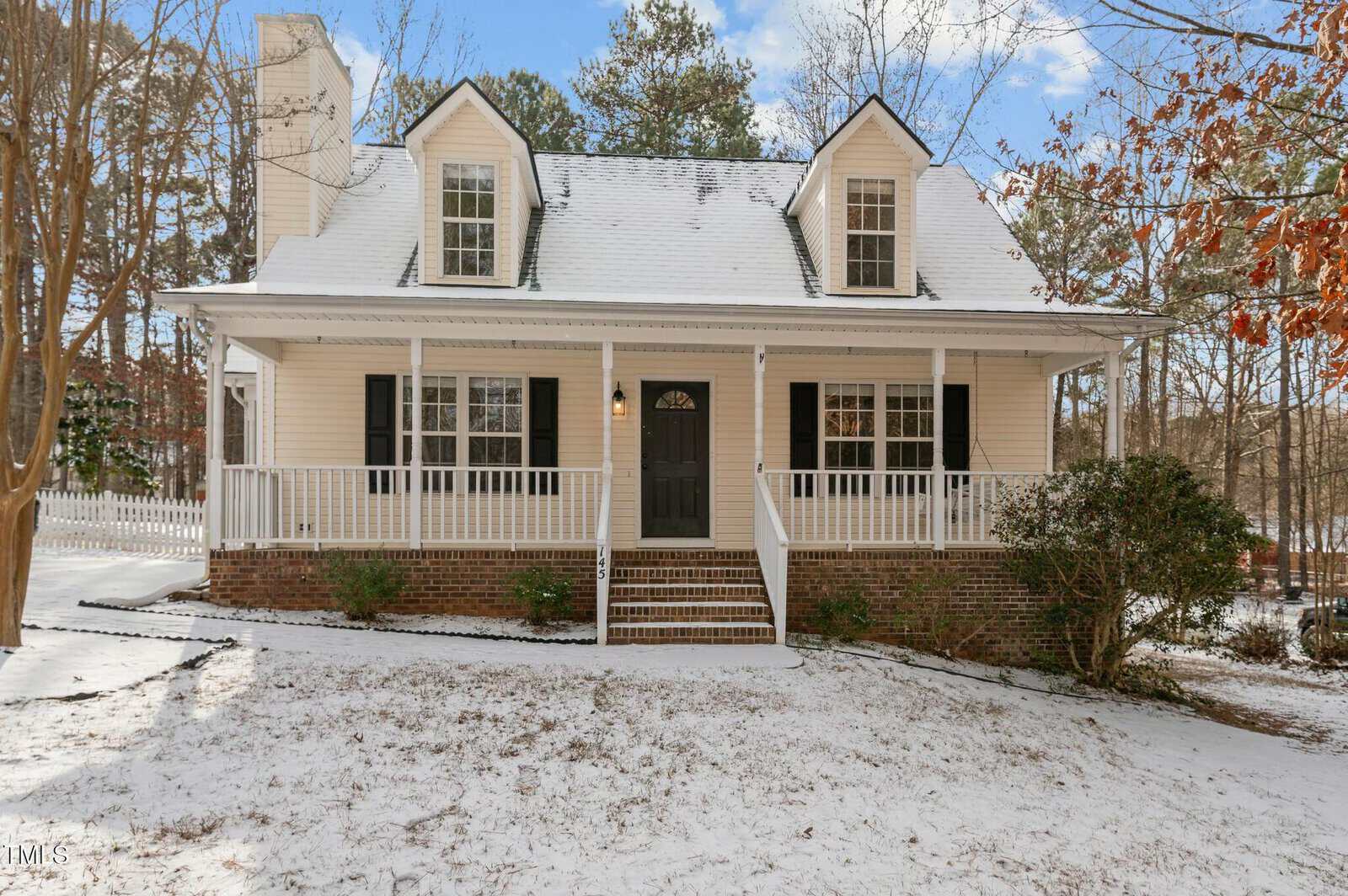 A two-story house with a covered front porch, black shutters, and a brick foundation. Snow covers the ground, roof, and porch, with trees and a blue sky in the background.