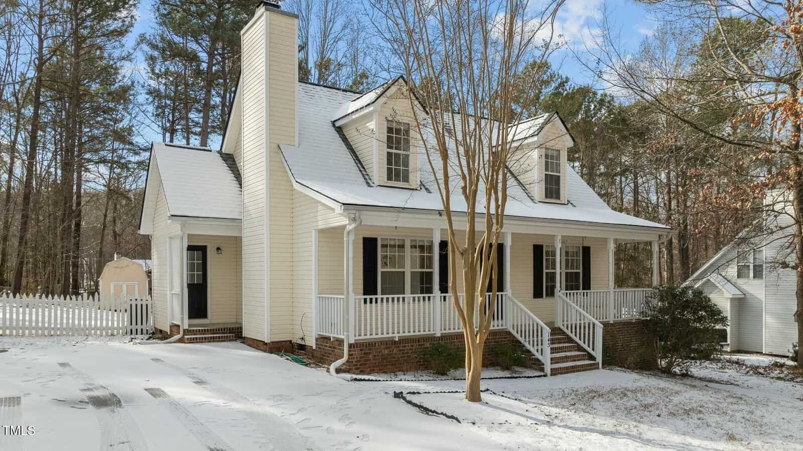 A light yellow two-story house with a front porch, white trim, and dormer windows is surrounded by snow on the driveway and yard. There are trees in the background and a white picket fence to the left.