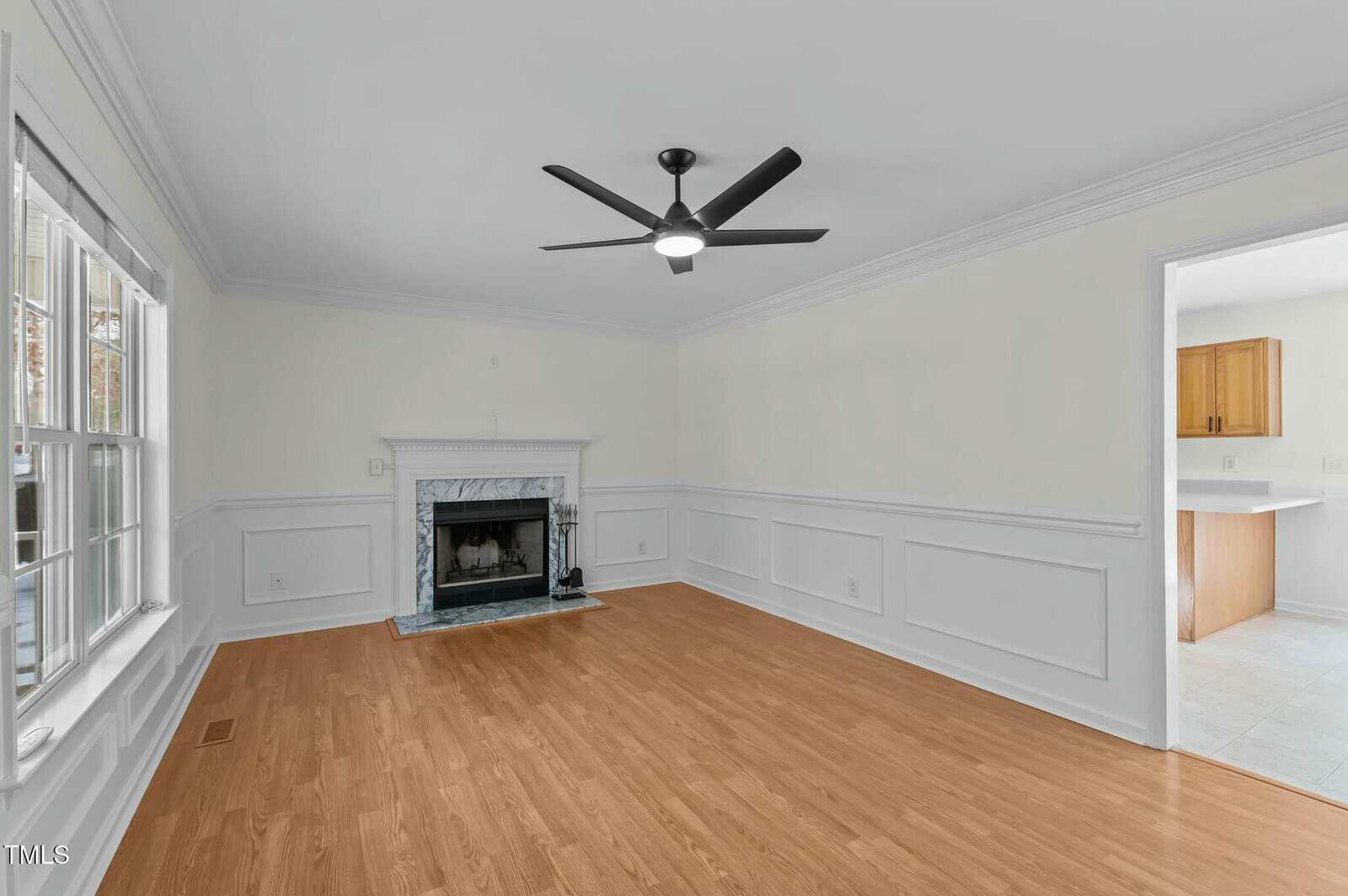 A bright living room with light-colored walls, hardwood floors, a black ceiling fan with light, a white fireplace, and large windows. An open doorway leads to a kitchen area with light cabinets and tile flooring.
