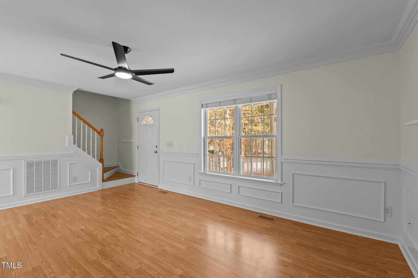 Bright living room with light wood flooring, white walls, large window, modern black ceiling fan, staircase with wooden railing, and front door. Natural light fills the space.