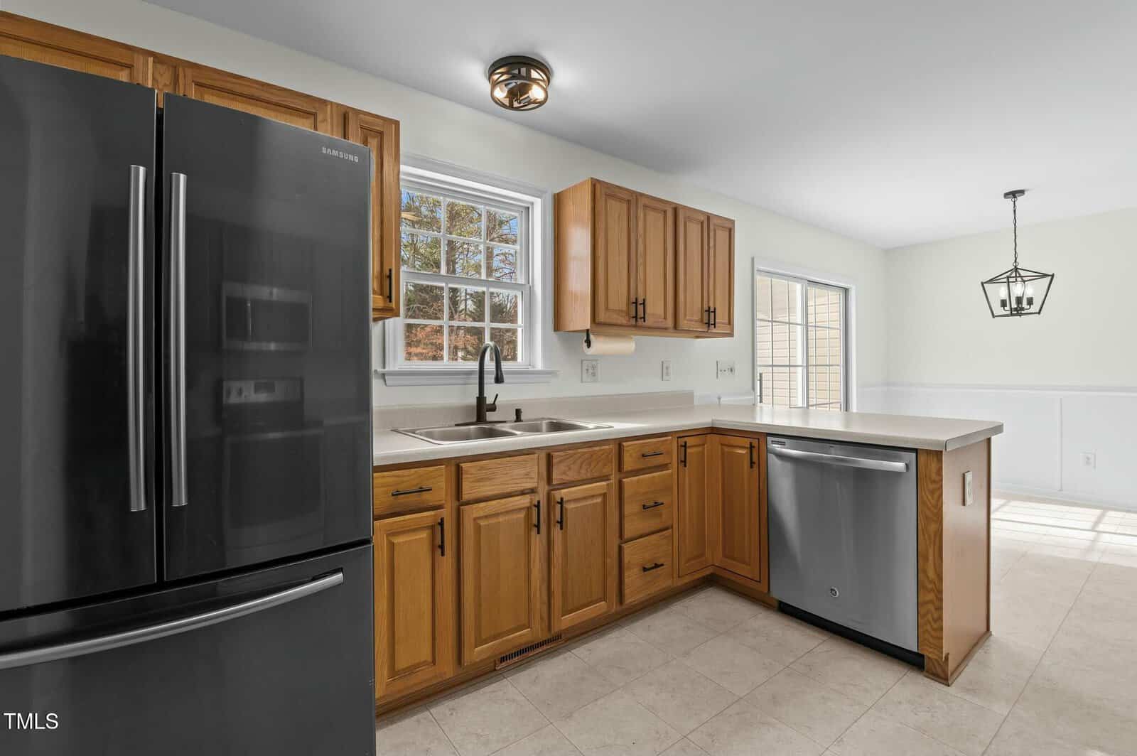 Kitchen with wooden cabinets, black refrigerator, stainless steel dishwasher, white countertops, and a window above the sink. A dining area with large windows and a modern light fixture is visible in the background.