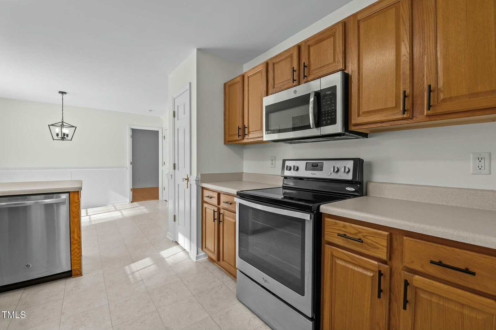 Modern kitchen with stainless steel appliances, light wood cabinets, beige countertops, and tile flooring. A windowed dining area with a hanging light fixture is visible in the background.
