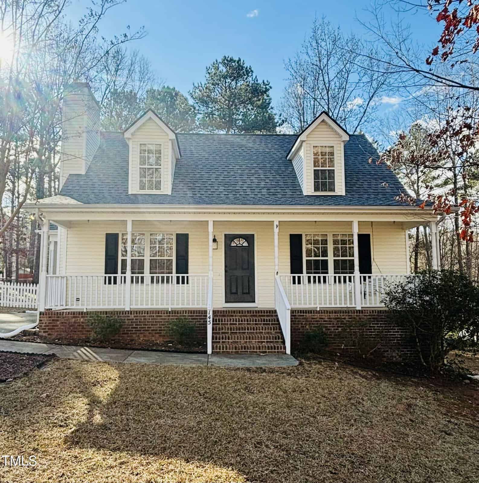 A two-story house with yellow siding, white trim, a large front porch with railings, two dormer windows, black shutters, and a dark front door. The front yard has brown grass and sparse trees in the background.