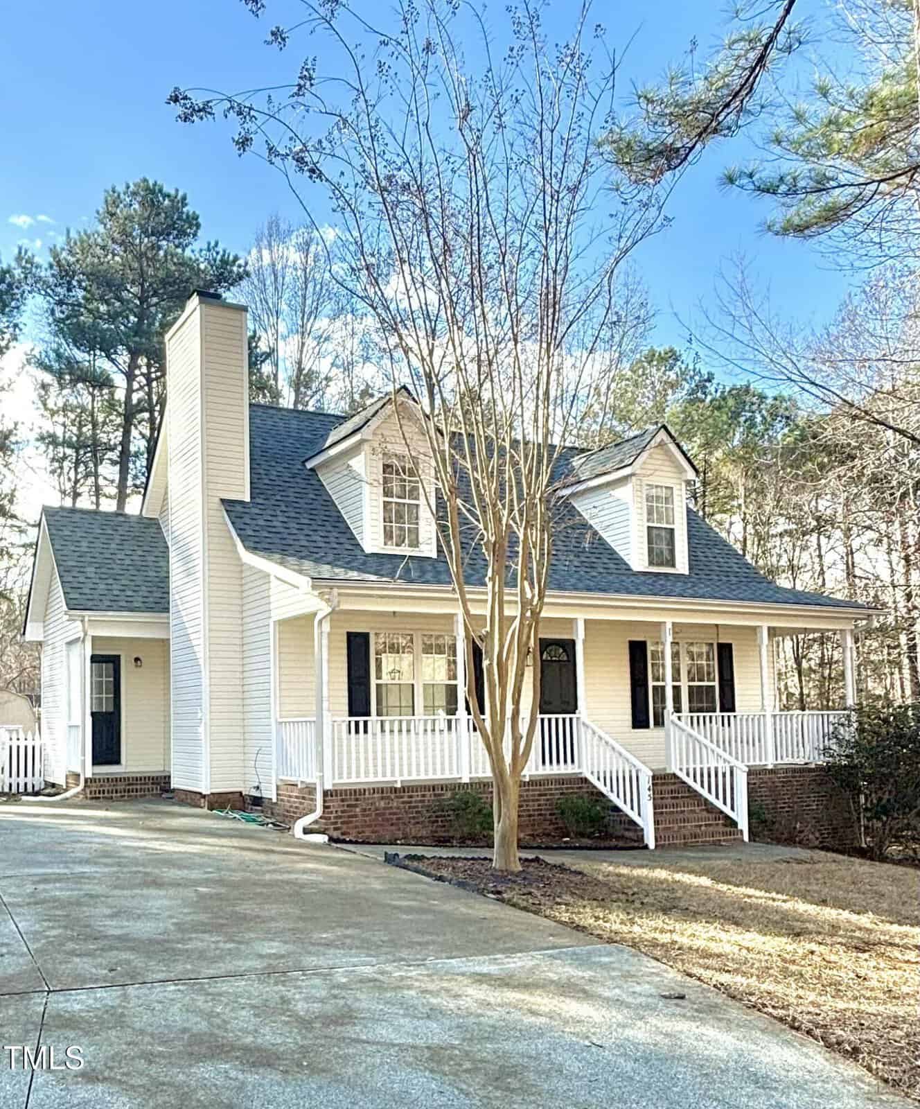 A two-story white house with a front porch, black shutters, and a gable roof. The house has a brick foundation, a chimney, and sits next to a paved driveway with surrounding trees and clear sky.