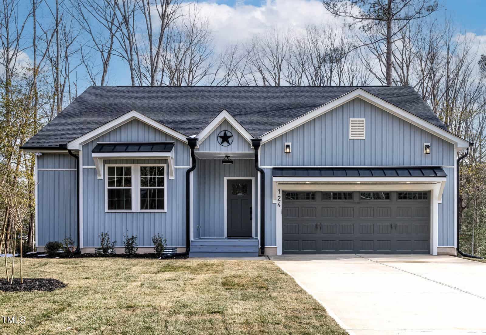 A single-story blue house with white trim, a front porch, a star decoration above the door, two front windows, a large gray double garage door, and a concrete driveway, surrounded by a grassy yard and trees.