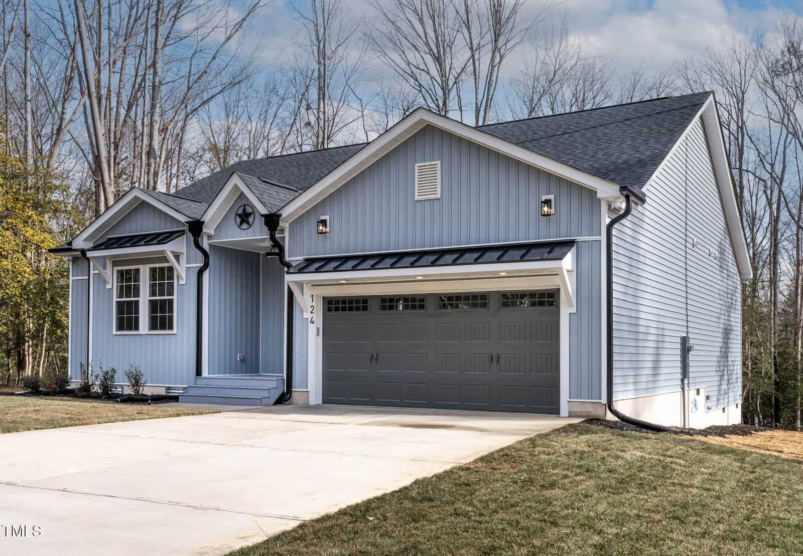 A light blue, single-story house with dark gray roof and attached two-car garage, set against leafless trees. The driveway is concrete and the lawn is small and neatly trimmed.