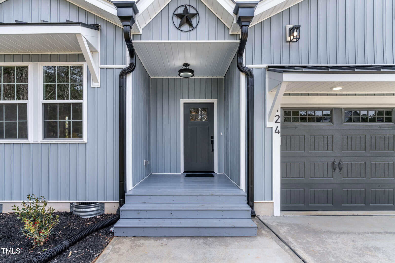Front entryway of a house with gray vertical siding, a covered porch with steps, a gray front door with a glass panel, a black star decoration above the door, and part of a garage door visible on the right.