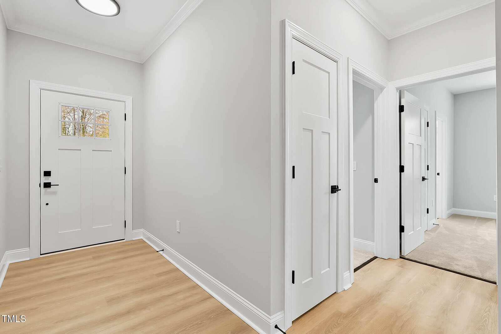 Bright hallway with light wood floors, white doors with black hardware, neutral gray walls, and crown molding. Multiple doors lead to different rooms; natural light comes in from a window on the door.