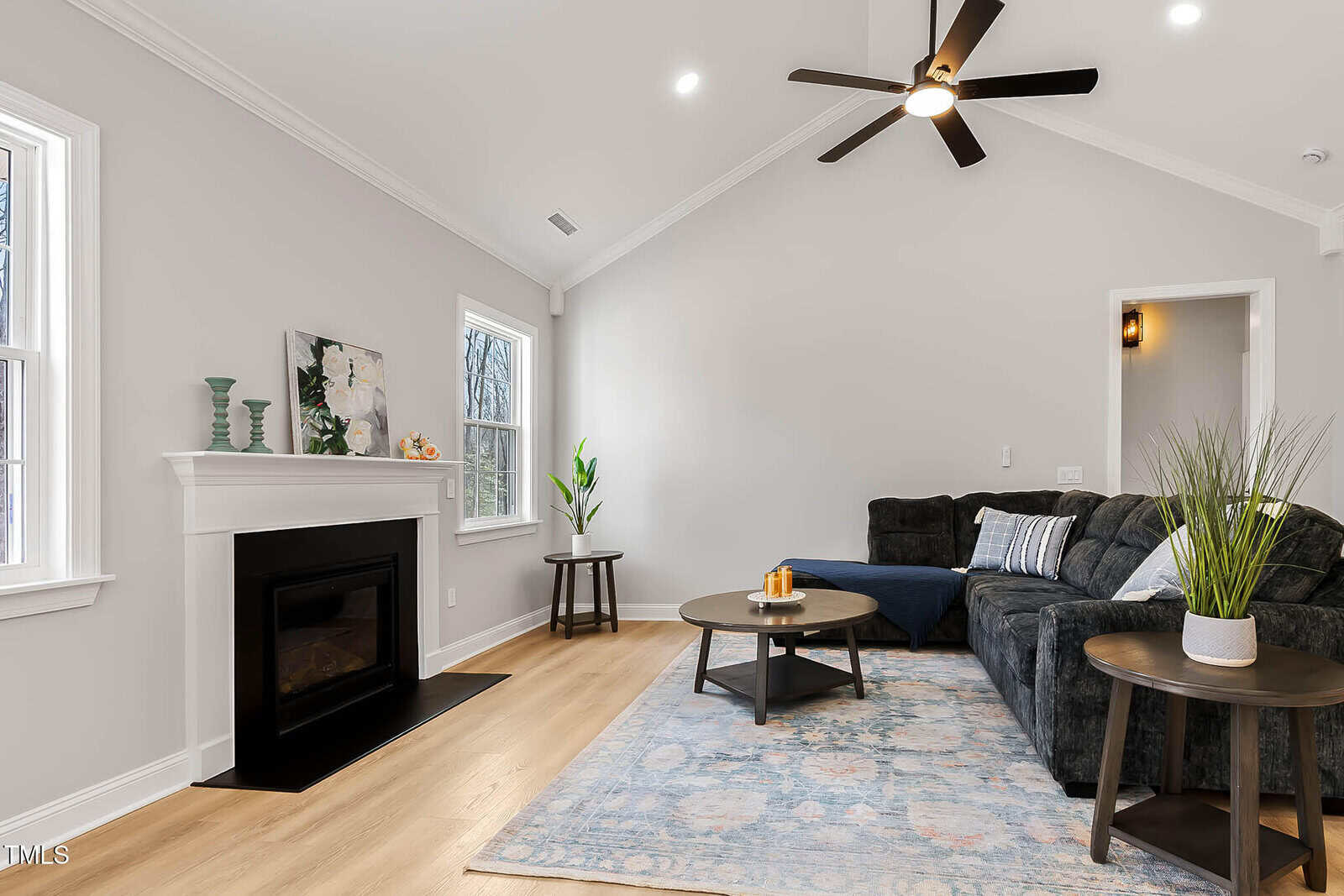 A modern living room with light gray walls, a black ceiling fan, a dark sectional sofa, a patterned rug, two round coffee tables, a fireplace with decor, and large windows letting in natural light.