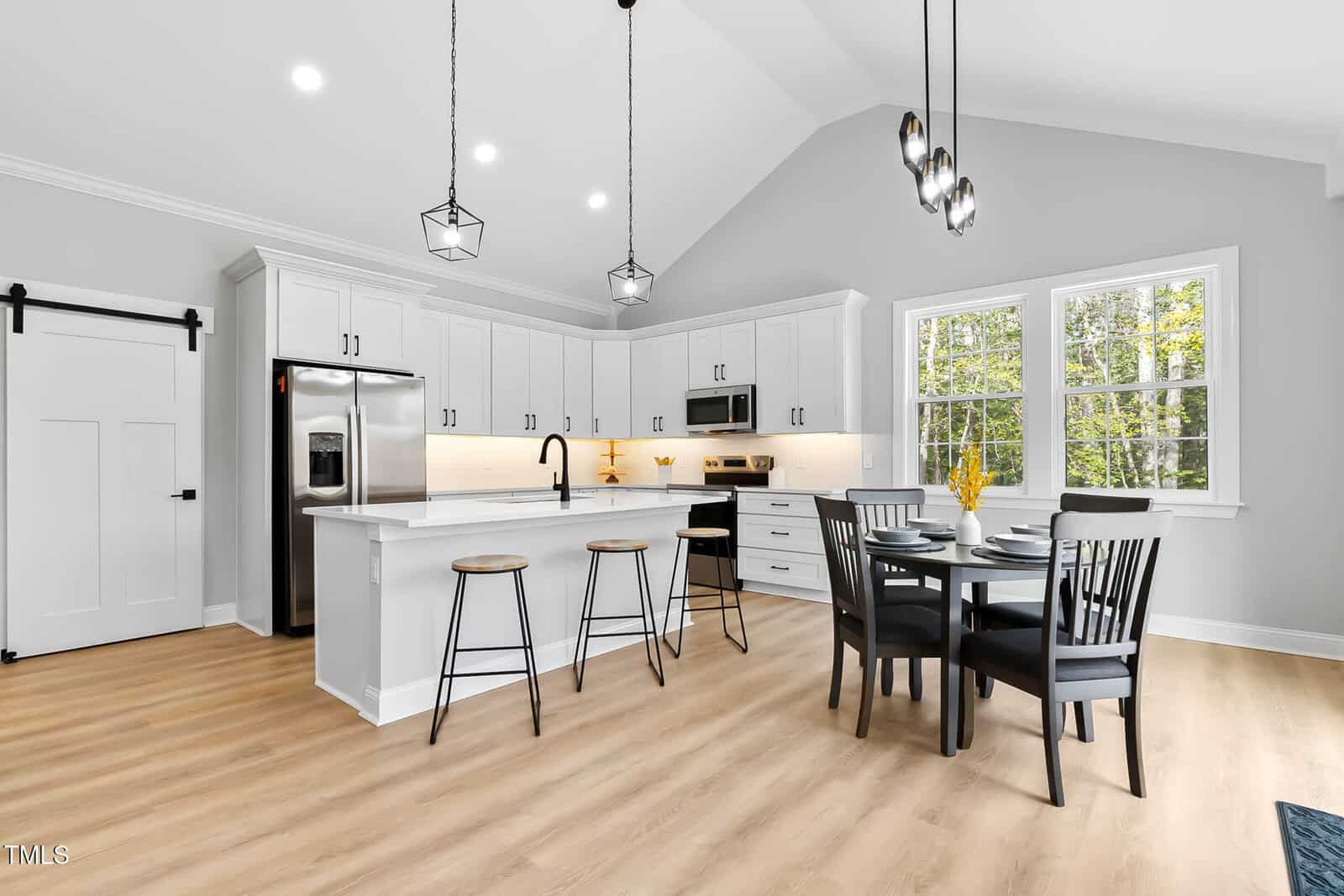 Modern kitchen with white cabinets, stainless steel appliances, an island with two bar stools, a round dining table with four black chairs, light wood flooring, and large windows letting in natural light.