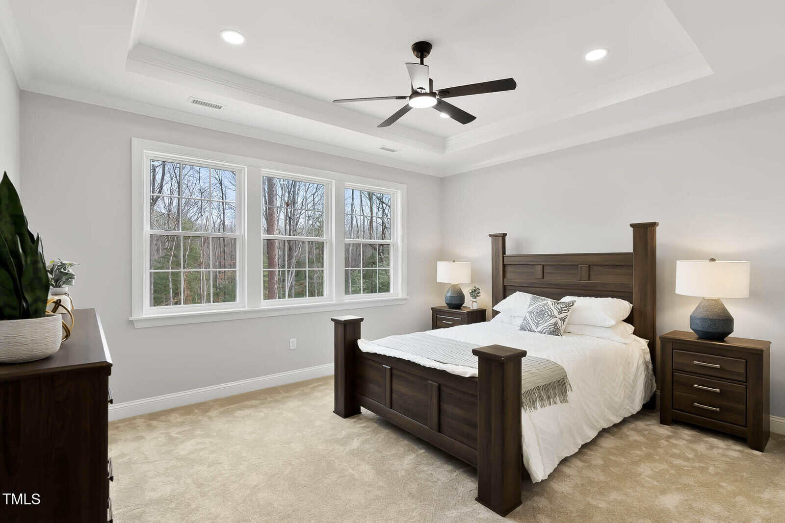 A modern bedroom with a wooden bed, matching nightstands, two lamps, a dresser with plants, beige carpet, white walls, and large windows showing a view of bare trees.
