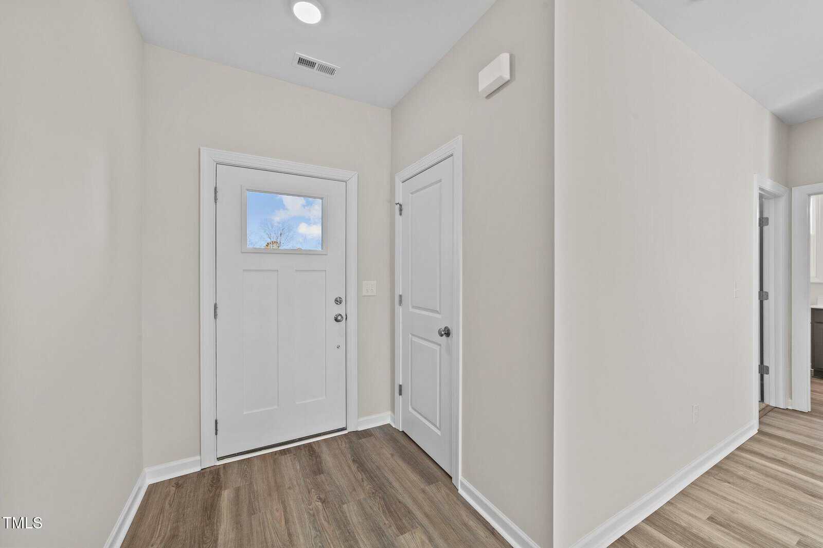 A hallway with a white front door featuring a small window, two white interior doors, beige walls, and wood-style flooring under ceiling lights.