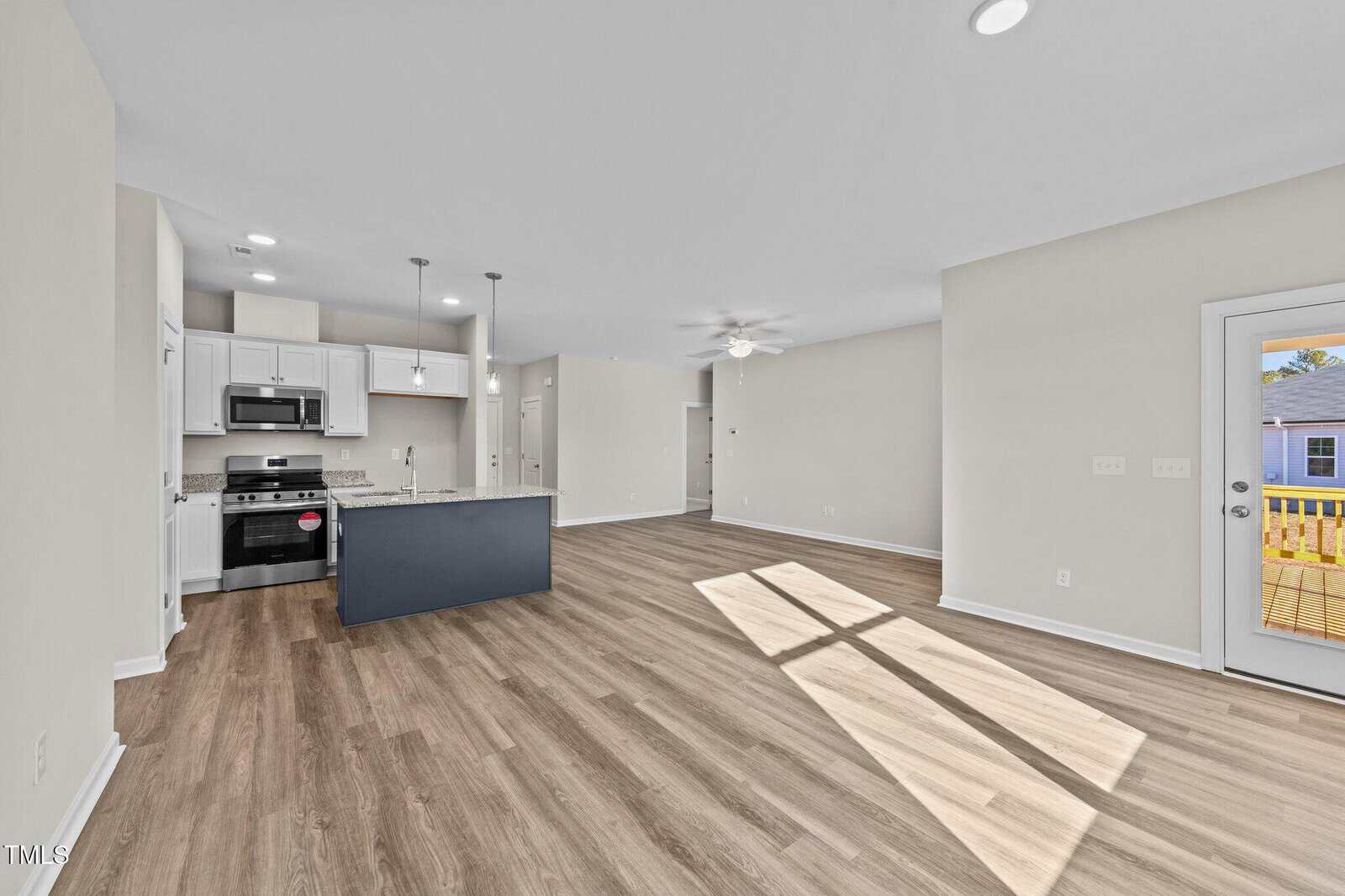 Open-concept living area with light wood flooring, white walls, and a modern kitchen featuring stainless steel appliances and a center island. A door on the right leads to an outdoor deck. Ceiling fan and recessed lighting are visible.