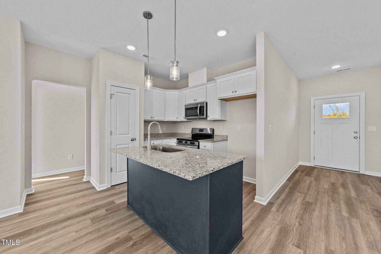 Modern kitchen with white cabinets, stainless steel appliances, and granite countertops. An island with a sink is centered under two pendant lights. The space has wood-style flooring and neutral-colored walls.