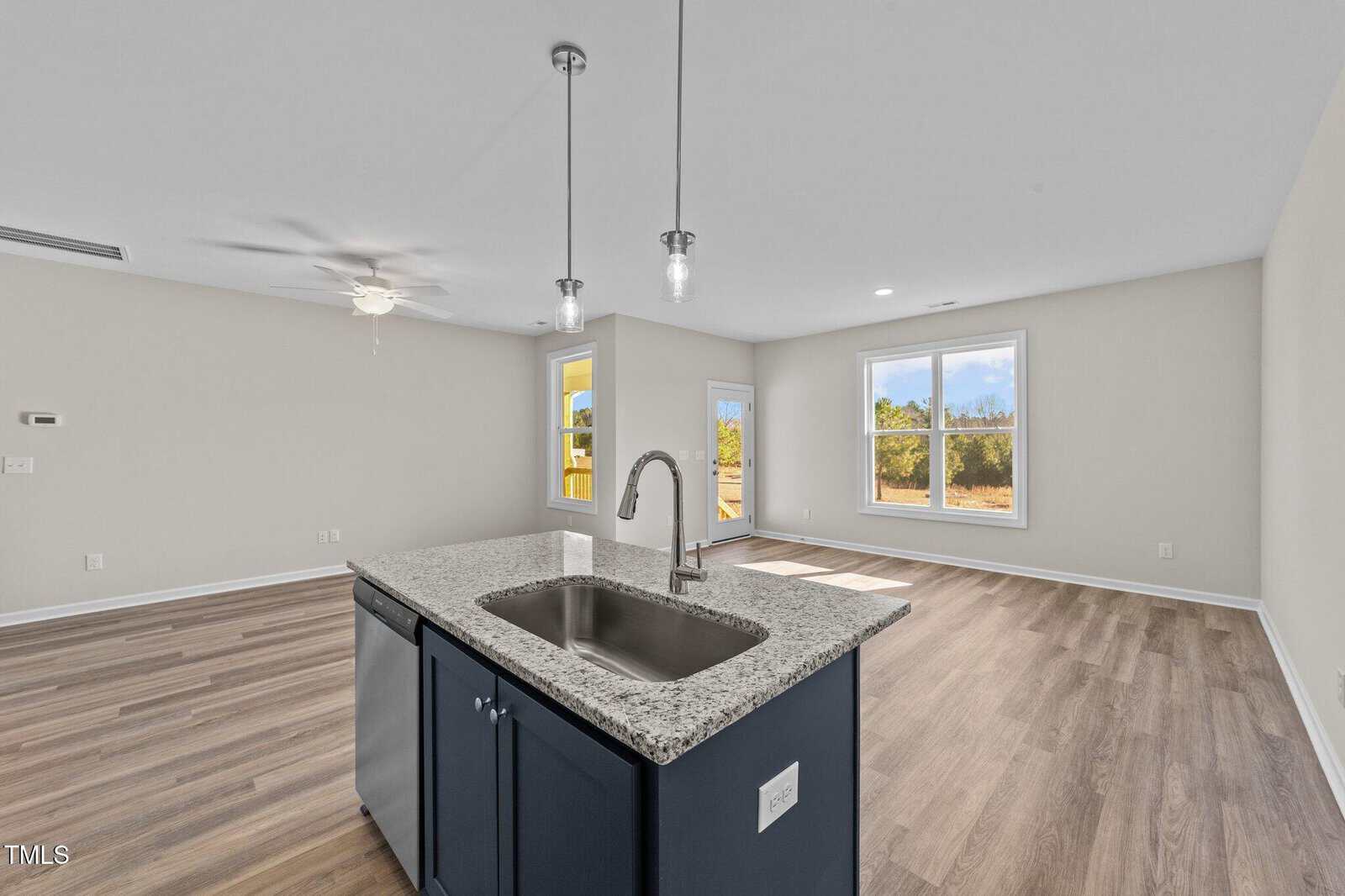 Open-plan living area with light wood flooring, neutral walls, a kitchen island with a granite countertop and sink, pendant lighting, ceiling fan, and large windows showing an outdoor view. Room appears empty and unfurnished.