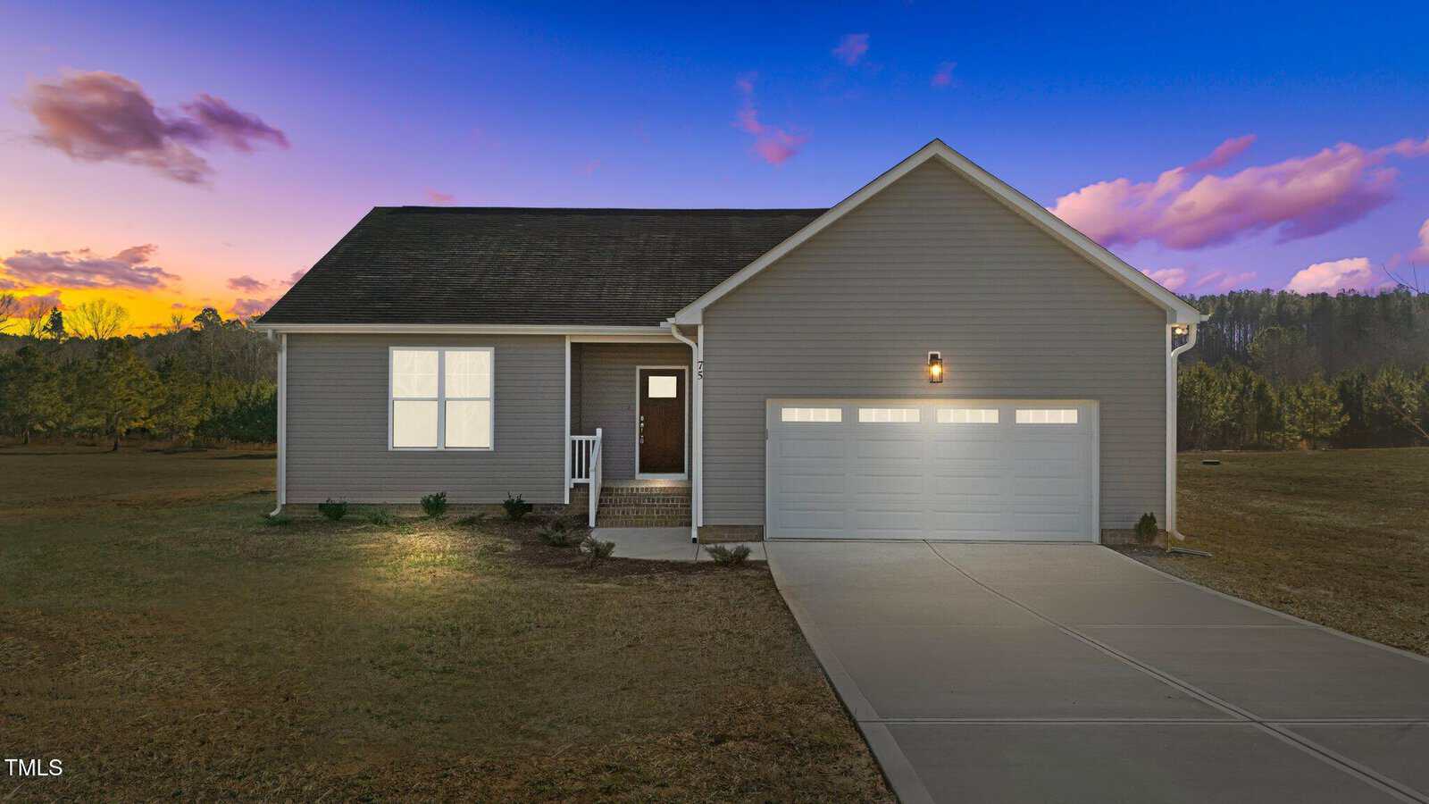 A single-story house with gray siding, a dark roof, and a two-car garage is pictured at sunset. The house has a small front porch, a concrete driveway, minimal landscaping, and a large open yard.