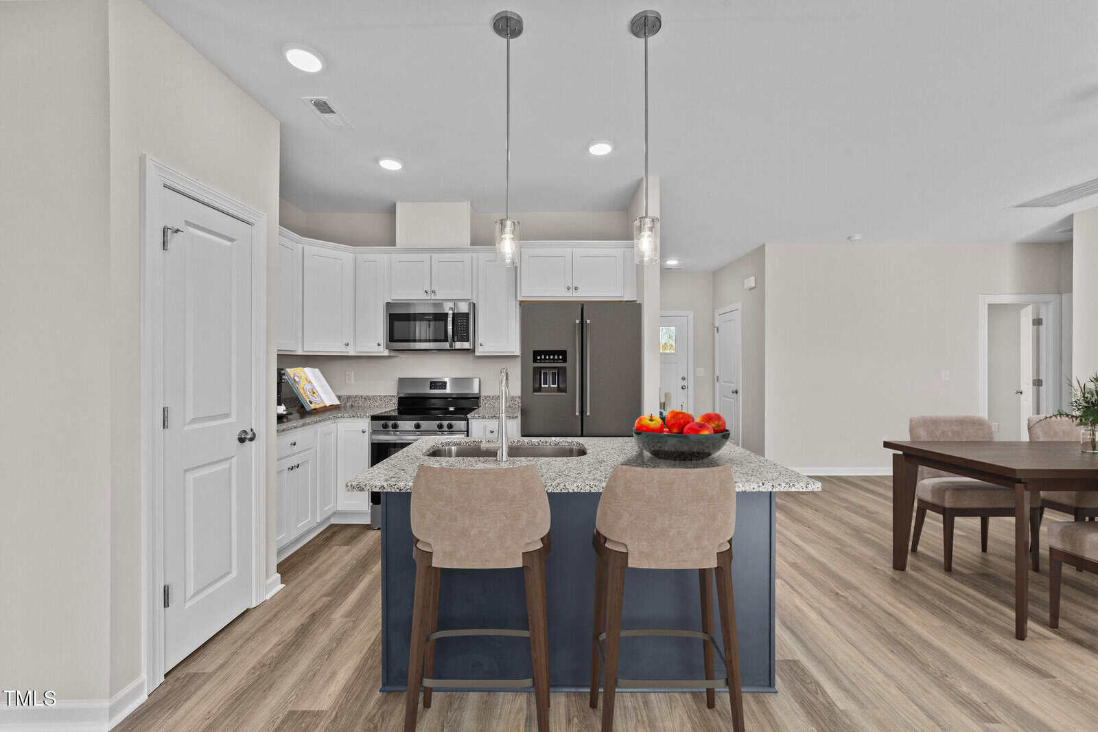 Modern kitchen with white cabinets, stainless steel appliances, a gray island with two beige chairs, pendant lights, and a bowl of apples. Dining area with a wooden table is visible in the background.