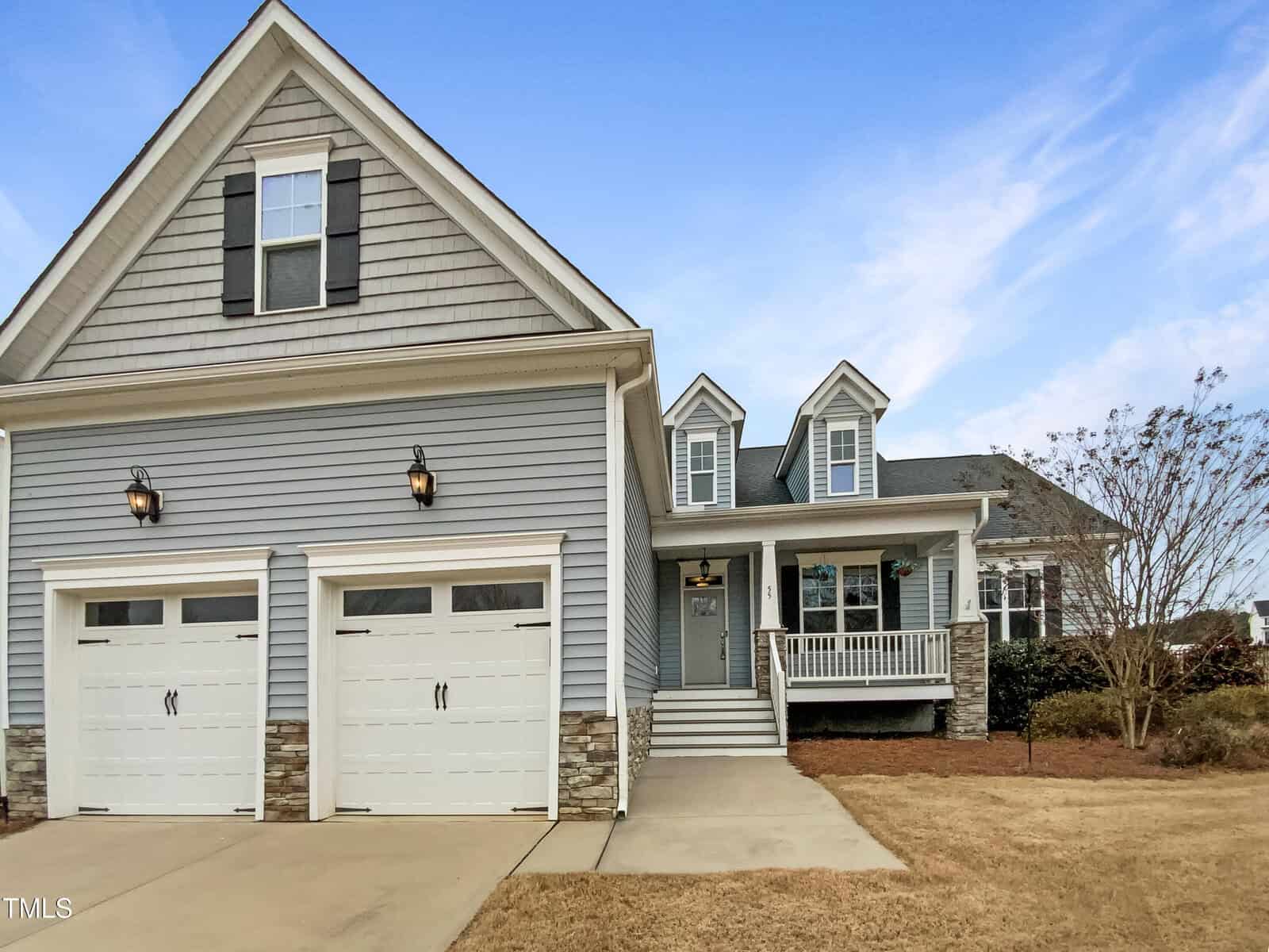A two-story suburban house with gray siding, two white garage doors, stone accents, dormer windows, a front porch with white railings, and a manicured lawn under a blue sky.
