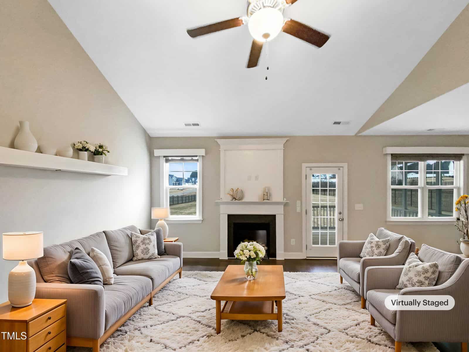 A living room with two gray sofas, a wooden coffee table with a flower vase, a fireplace, lamps on side tables, and a ceiling fan. Decor includes plants and vases; a rug covers the floor. "Virtually Staged" text appears.