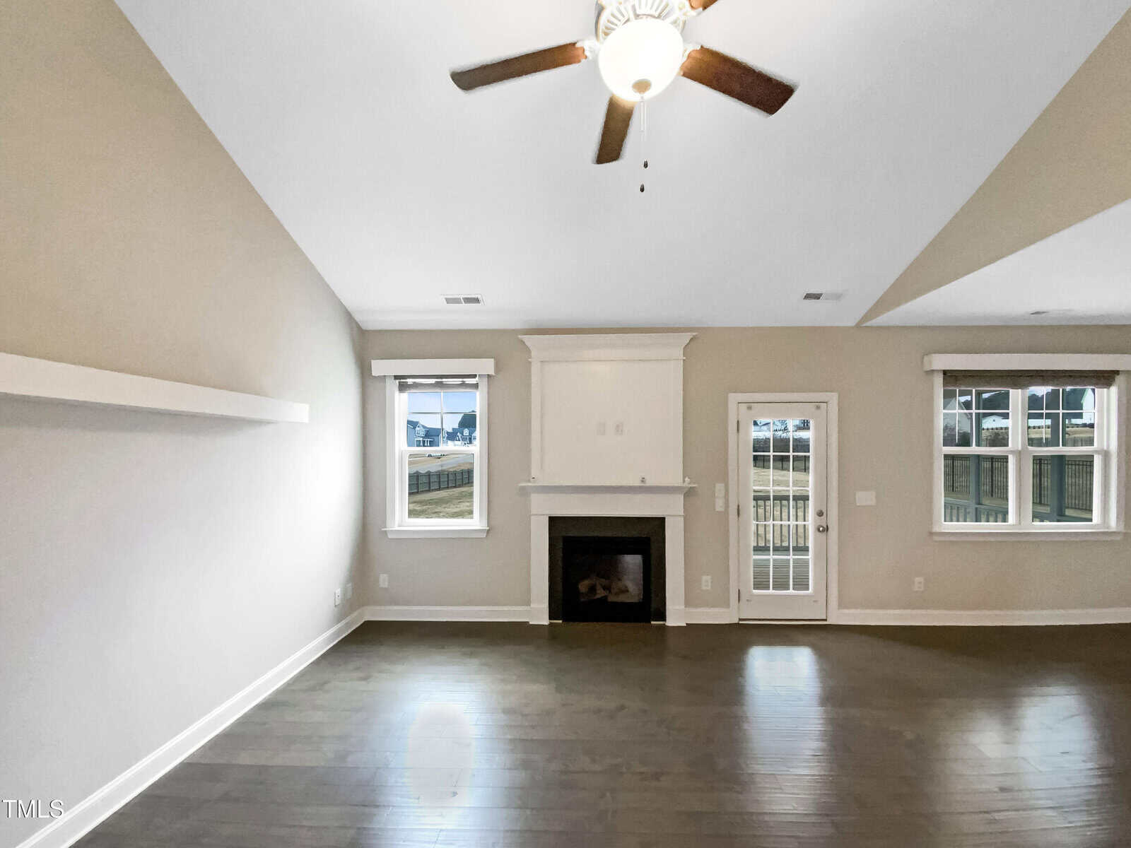 A spacious living room with light-colored walls, a ceiling fan, a fireplace centered between two windows, a glass door to the outside, dark wood flooring, and a floating wall shelf on the left.