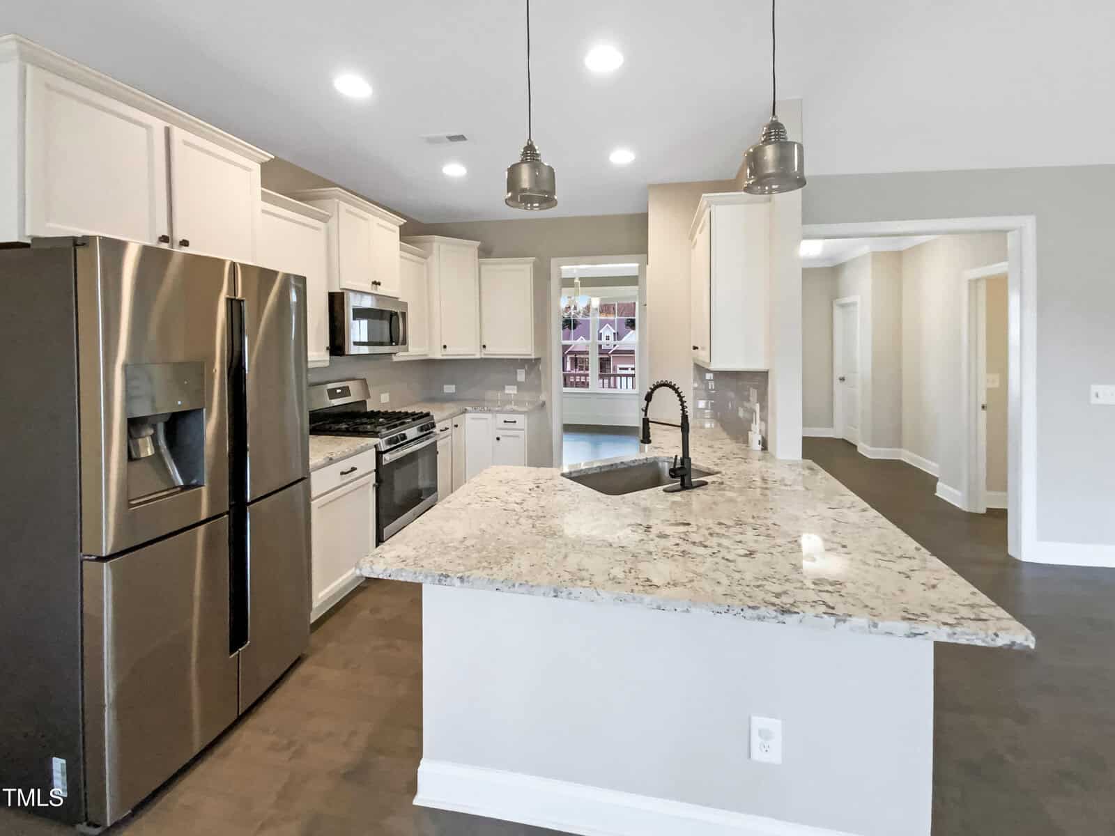 Modern kitchen with stainless steel appliances, white cabinets, granite island with sink, pendant lights above, and gray tile flooring. A hallway and an open doorway are visible in the background.