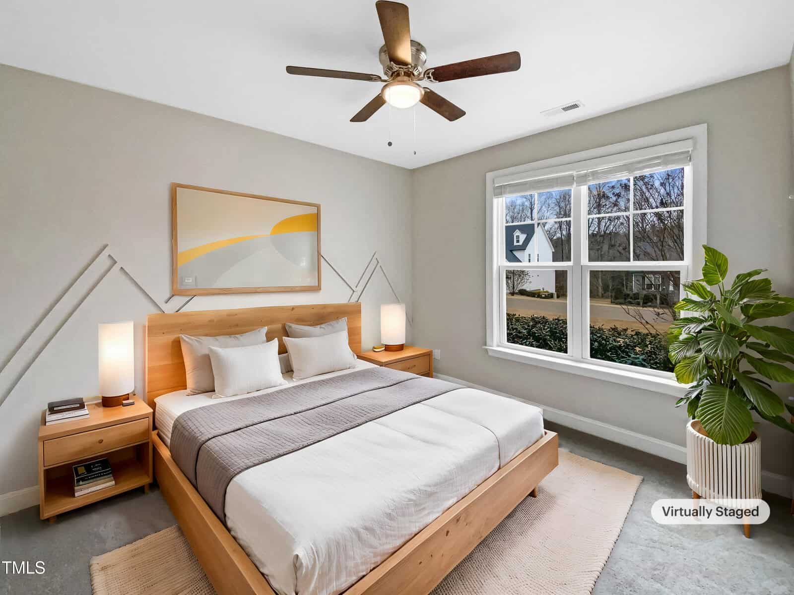 A bedroom with a wooden bed frame, two matching nightstands, white bedding with a gray blanket, abstract wall art, a ceiling fan, large window, and a potted plant; marked as virtually staged.