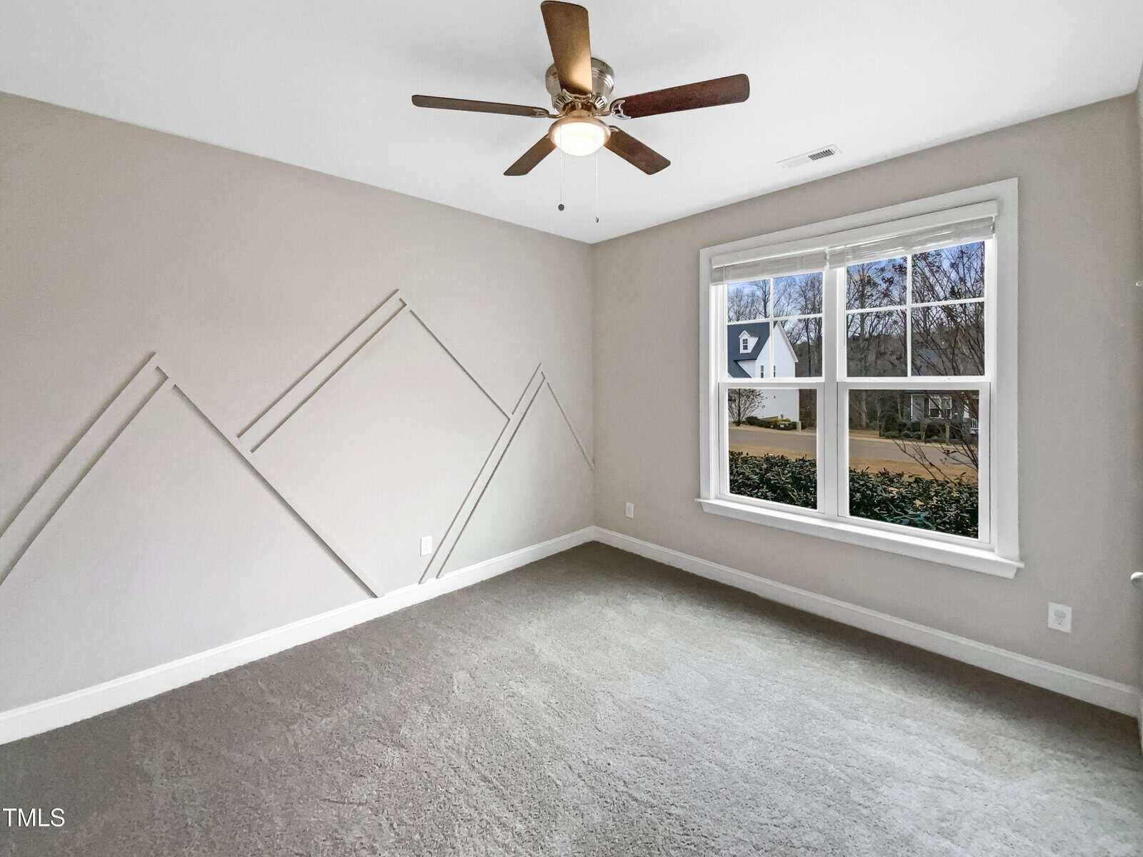 Empty room with gray carpet, light beige walls featuring angular decorative trim, a ceiling fan with light, and a large window showing a neighborhood street and trees outside.