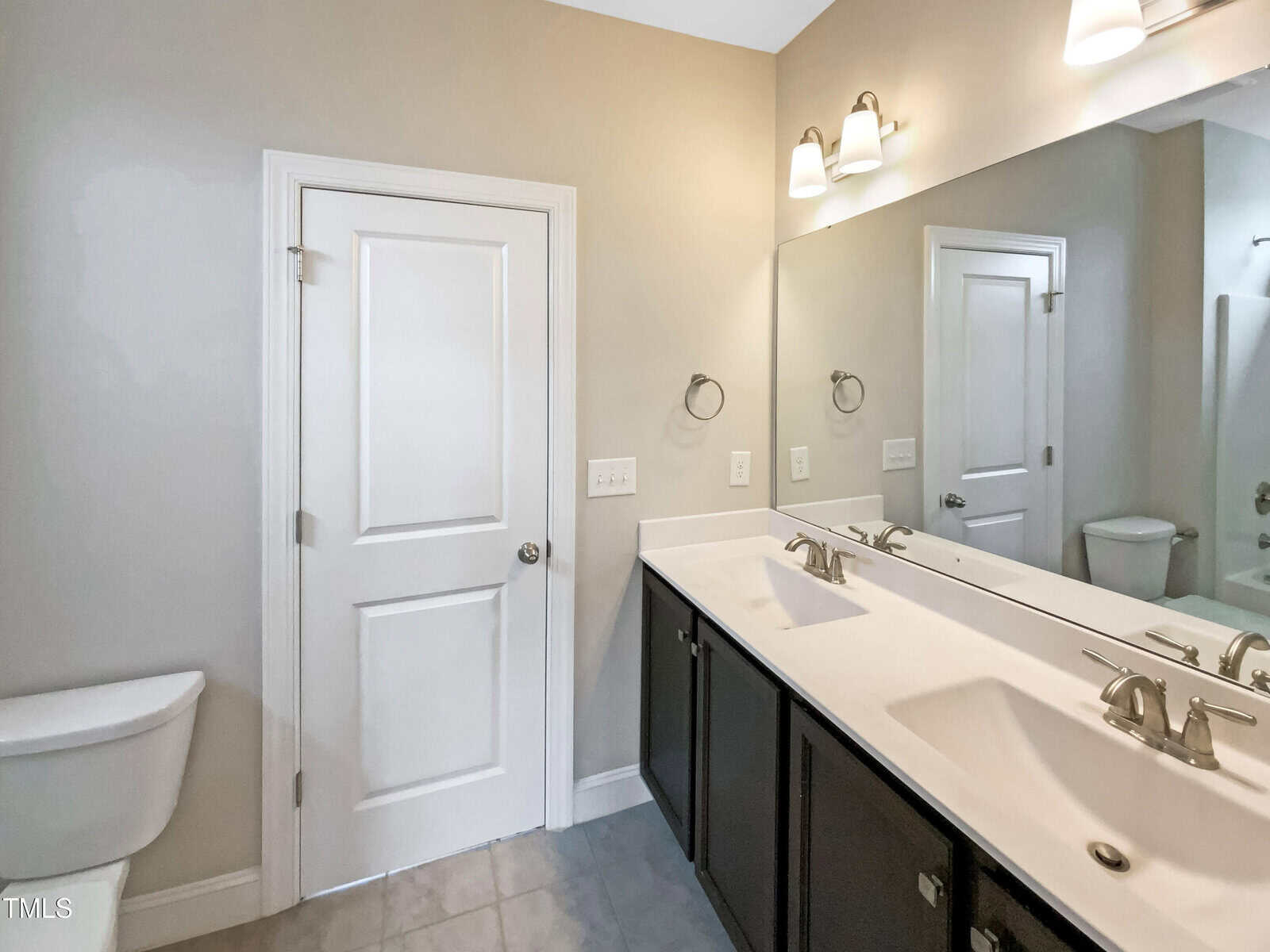 A bathroom with a double sink vanity, large mirror, two light fixtures, a white door, a toilet, and beige walls. The floor is tiled in a light color.