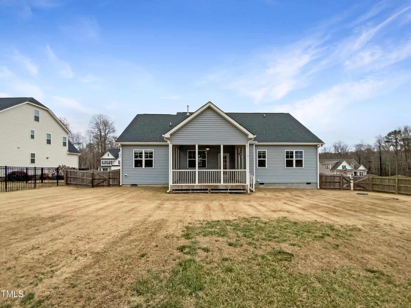 A single-story, light blue house with a covered back porch, white trim, and a large fenced backyard. The lawn is mostly brown with some green patches. Neighboring houses and trees are visible in the background.